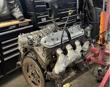 A V8 engine suspended by a crane in a garage, featuring silver valve covers and exhaust manifolds with heat shields.

