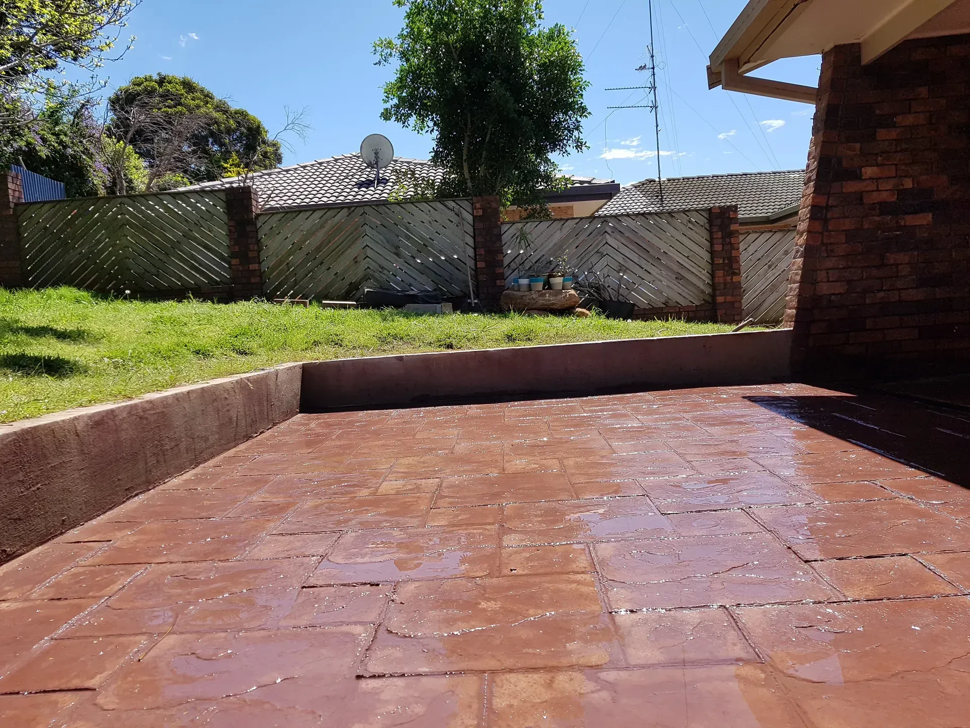 Patio with brick-colored pavers, surrounded by a low wall and a grassy yard. A fence and a tree are in the background.