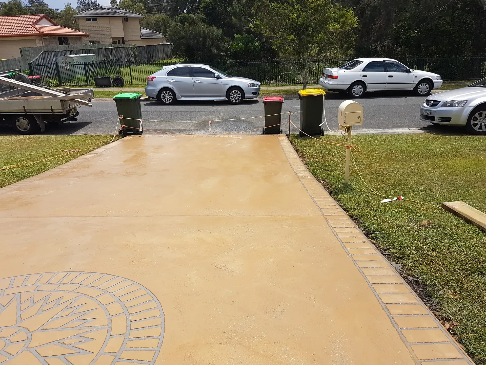 Driveway with trash bins, cars parked on street, and a house in the background on a sunny day.