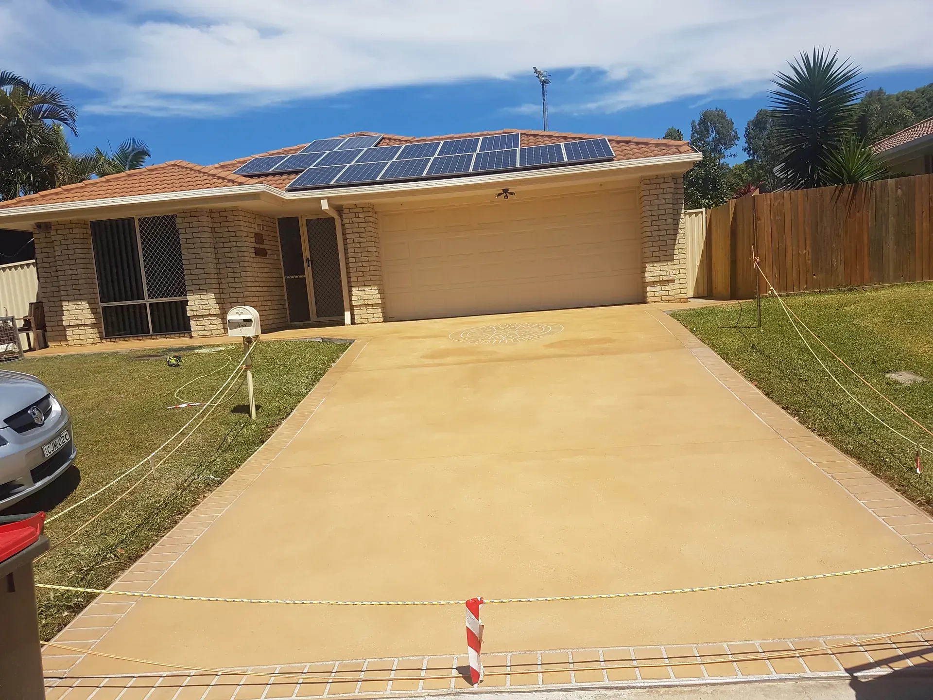 House with solar panels, tan driveway, and garage door. A car is parked on the left. Bright sunny day.
