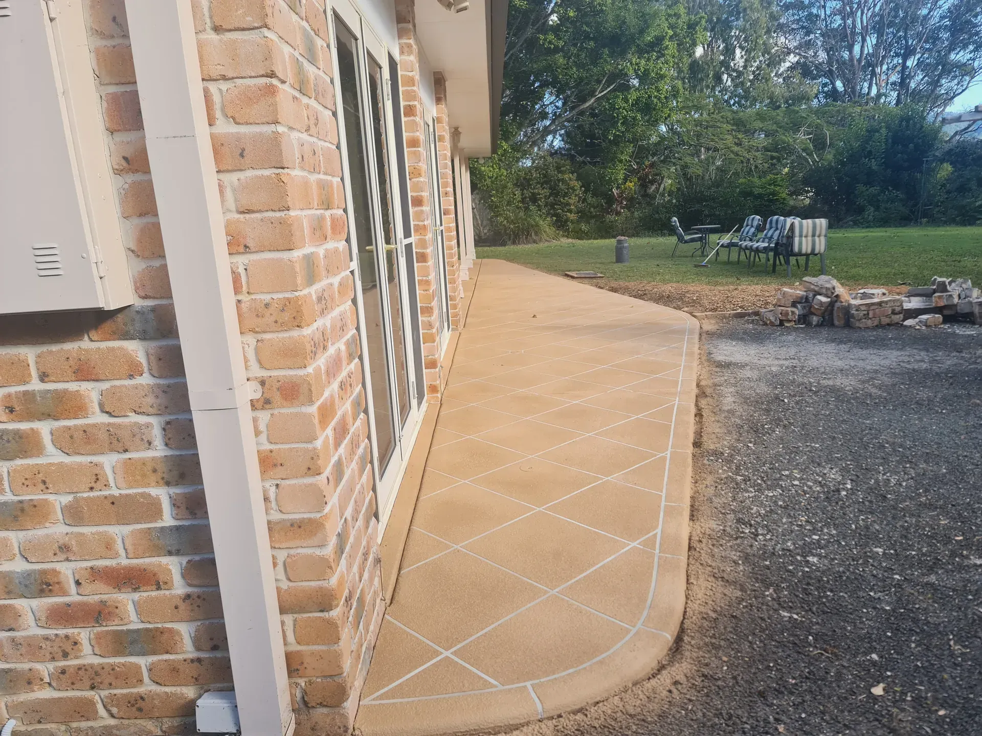 A concrete walkway curves along a brick building with windows. Gravel and trees are in the background.