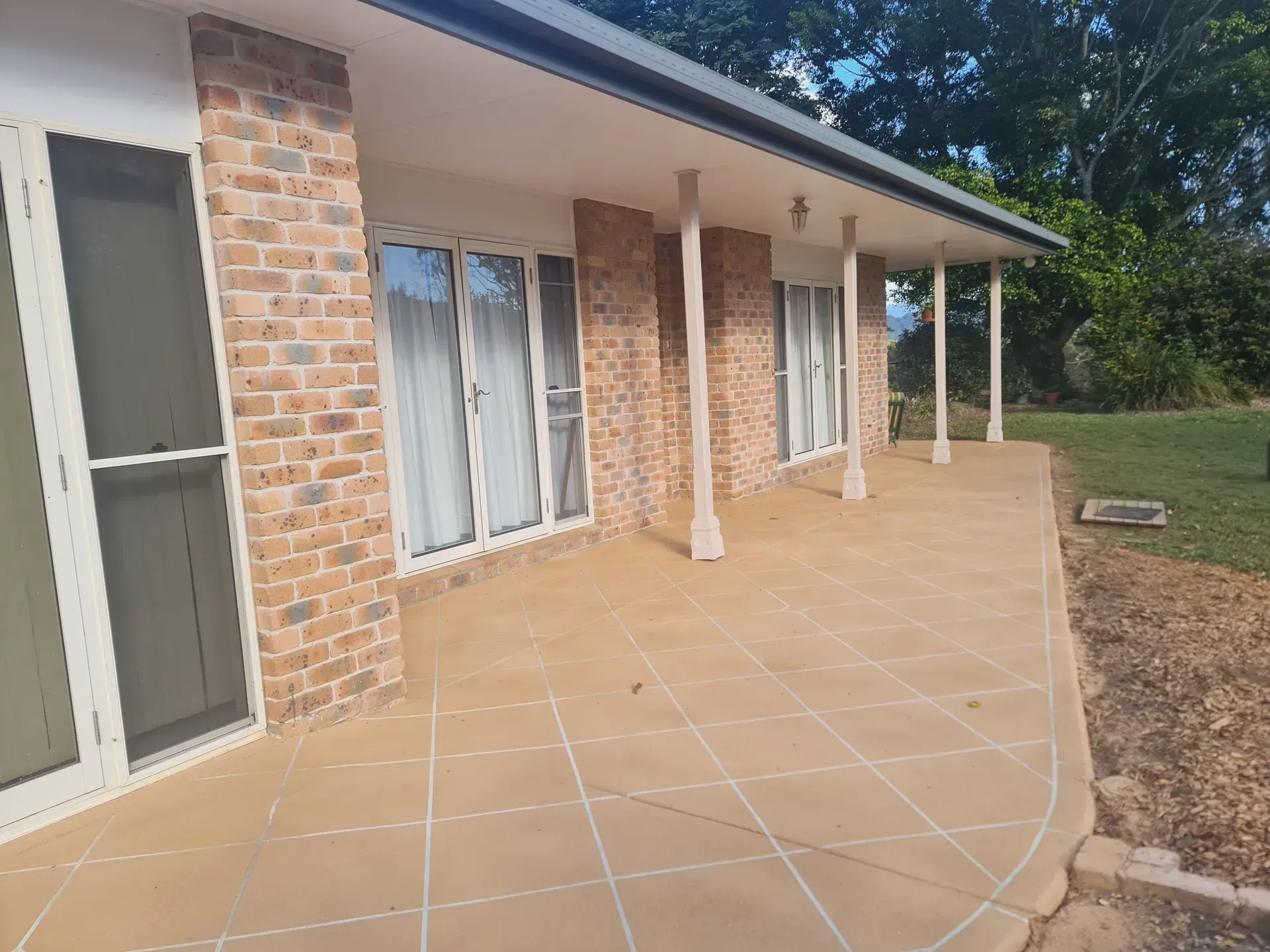 Exterior of a brick house with a covered patio. Beige tiles, white pillars, and large windows are visible.