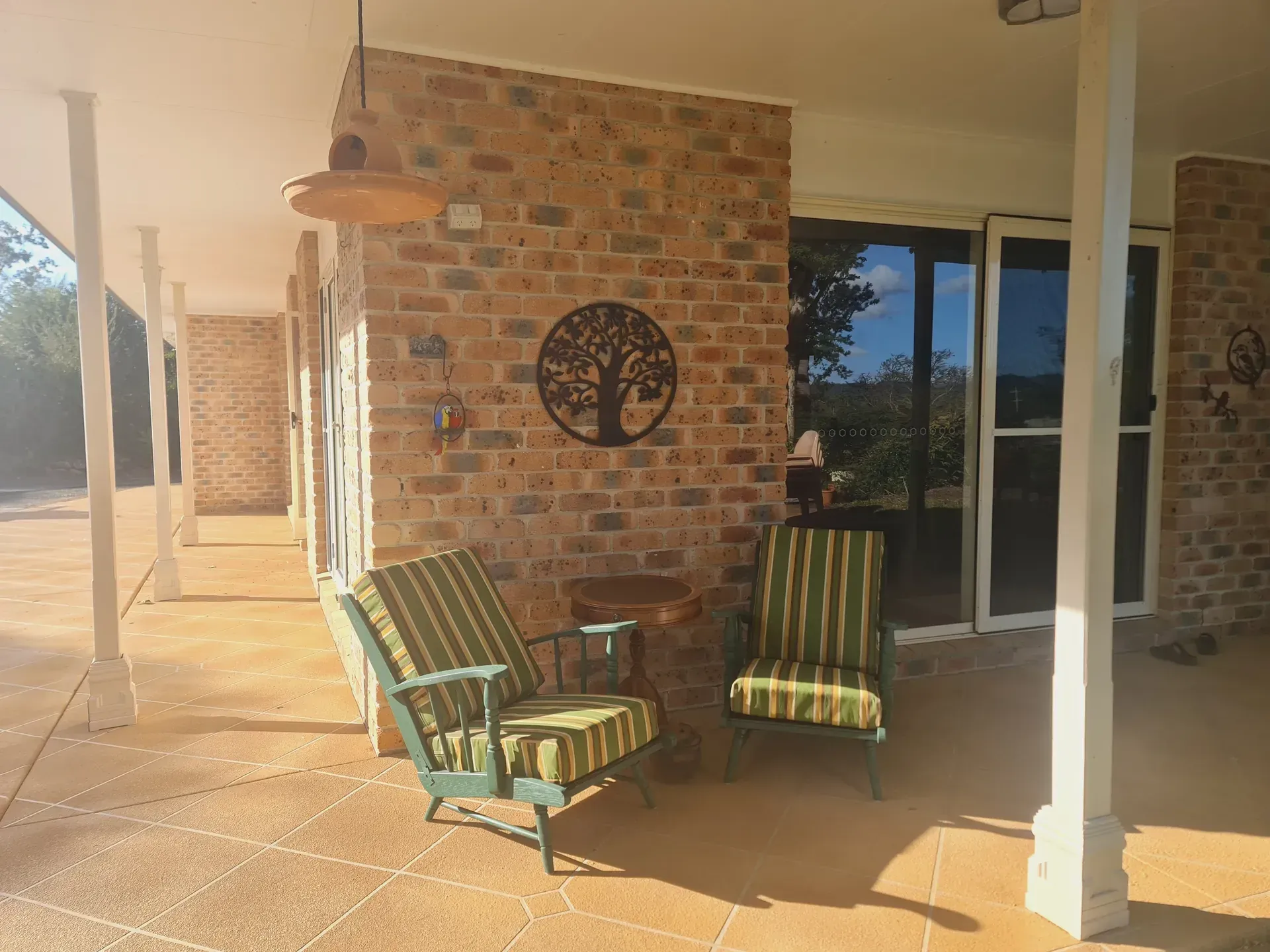Covered porch with brick wall, two striped chairs, and a metal tree decoration.