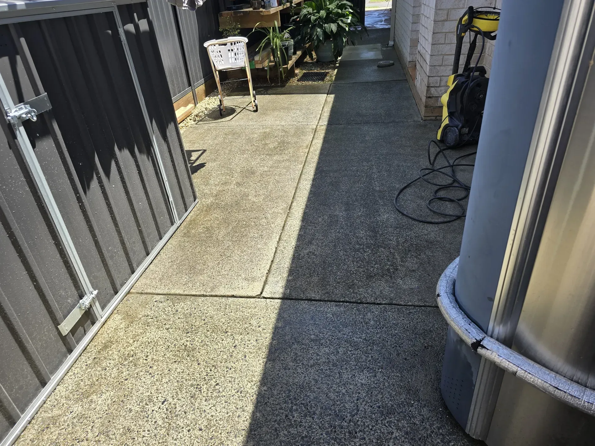 Concrete walkway between a gray fence and a building, with a pressure washer visible.