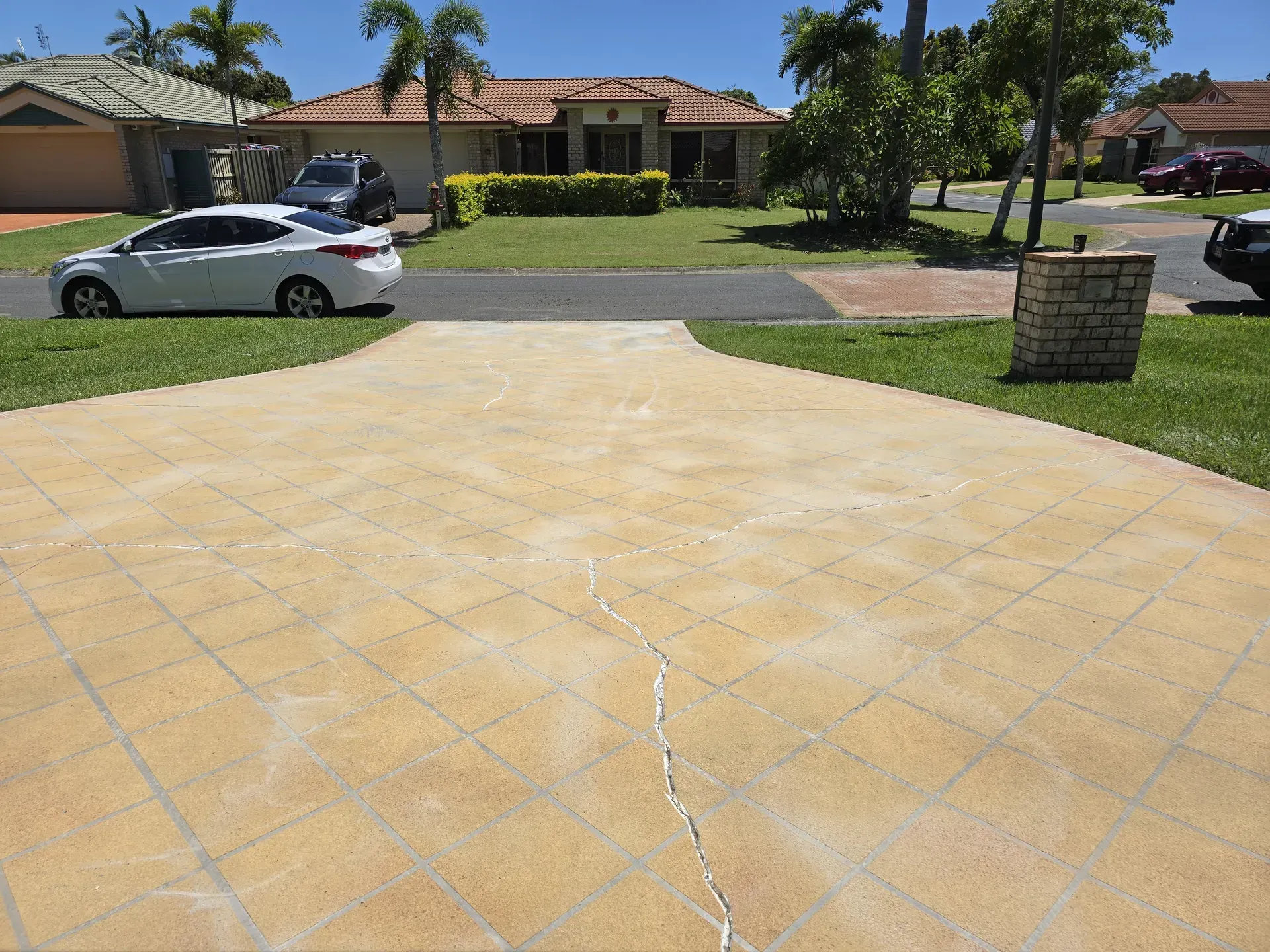 Cracked, yellow tiled driveway in front of a house, white car parked. Green lawn and sunny day.