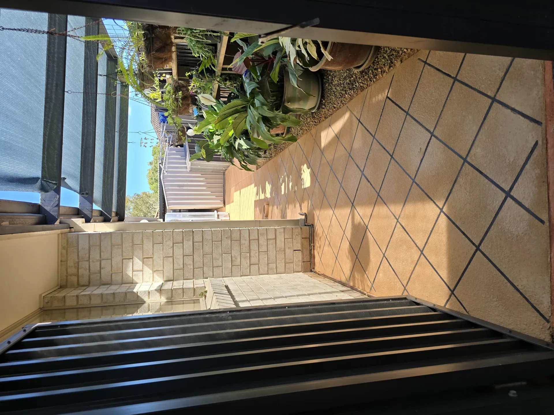 Staircase and patio with plants viewed from a doorway. Tiles are brown and plants are green. Bright sunlight.