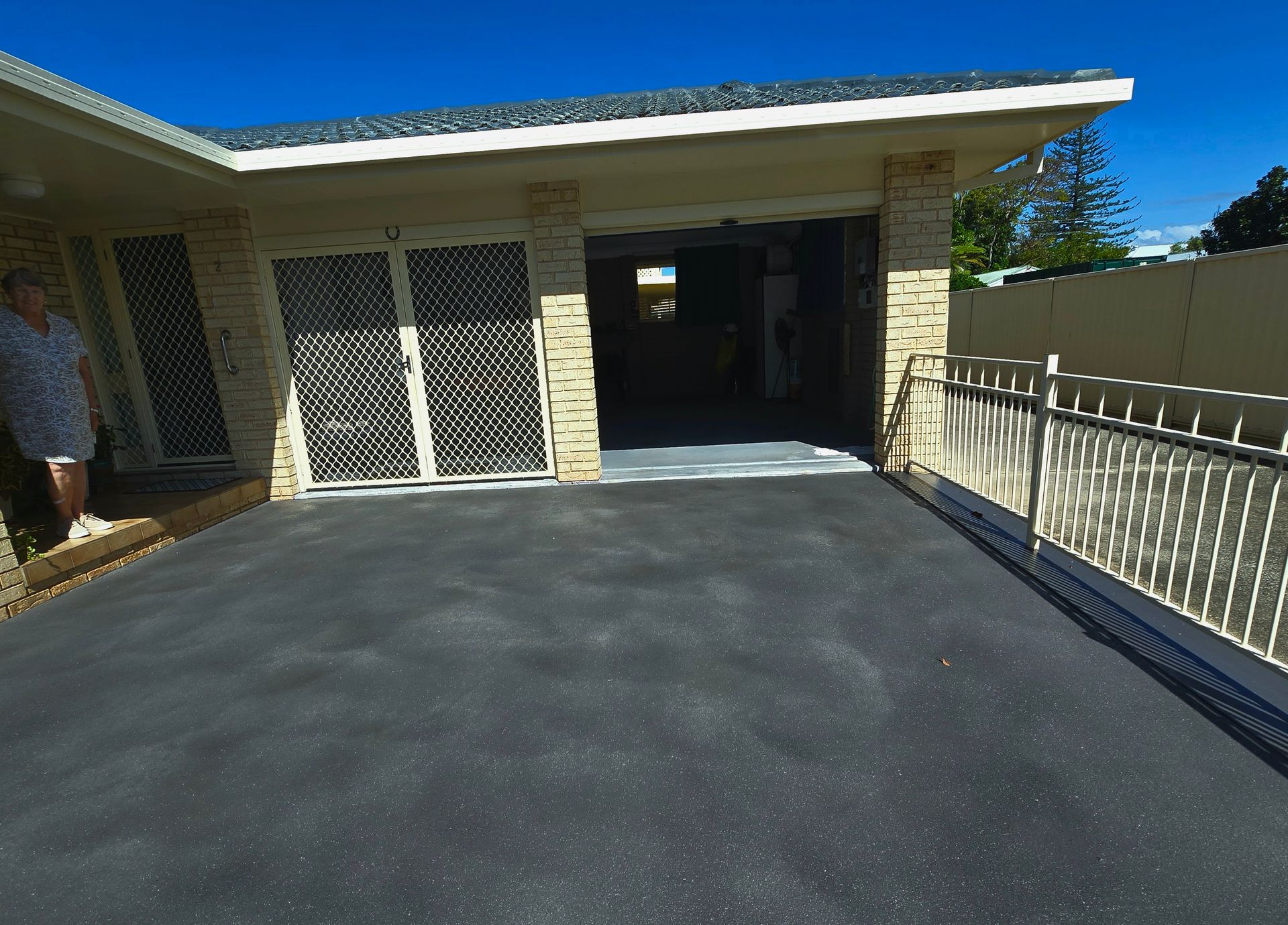 Driveway with open garage, woman standing at doorway. Dark asphalt, light tan brick, white security doors.