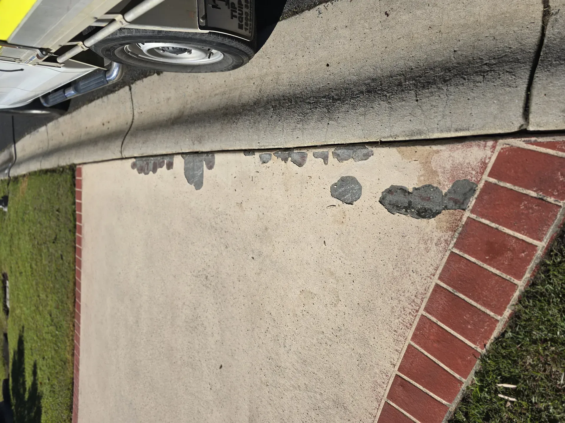 Damaged concrete sidewalk edge with visible patches near a curb and grass. Brick edging is also visible.