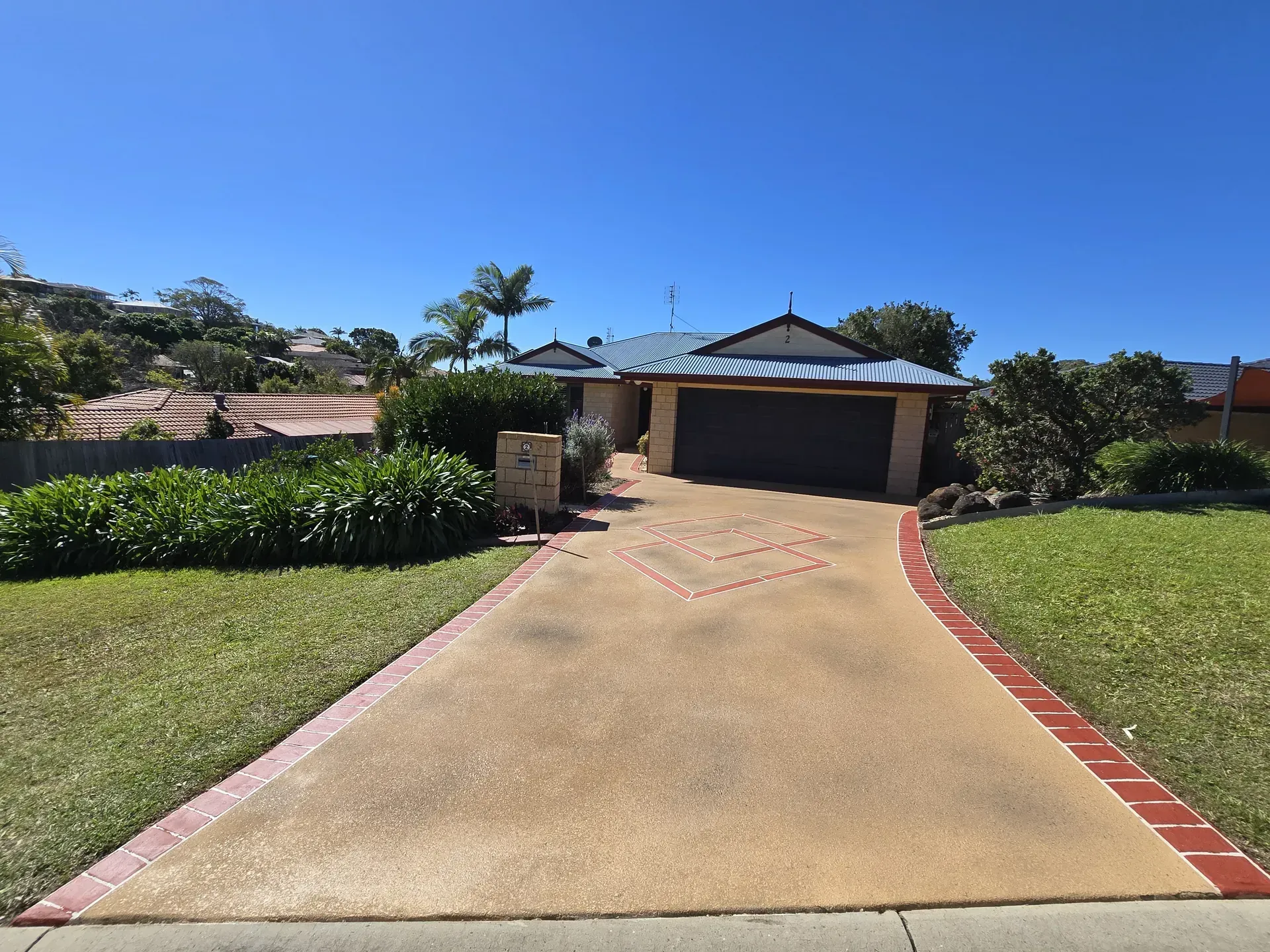 House with a tan driveway trimmed in red brick, a blue roof, and green lawn under a clear blue sky.
