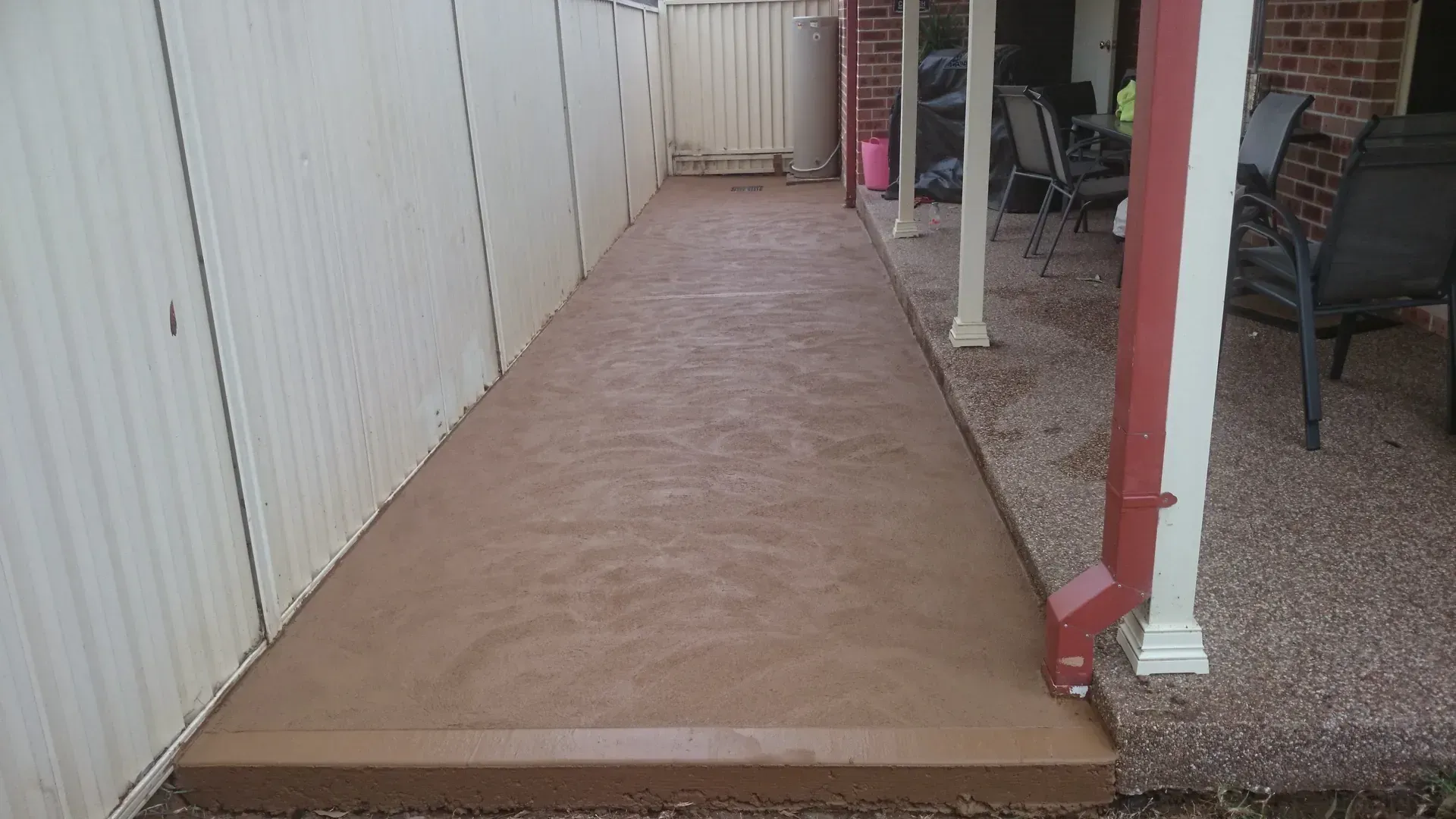 Brown concrete walkway between a white fence and a porch with chairs.