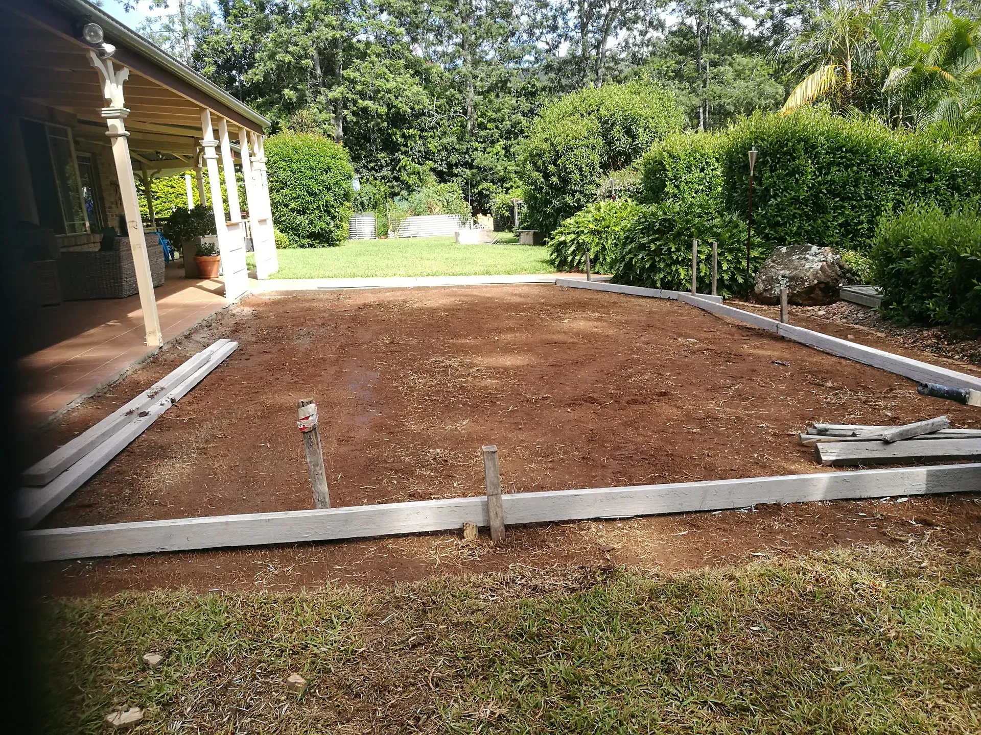 A rectangular area of red soil bordered by concrete forms, ready for construction in a grassy yard.