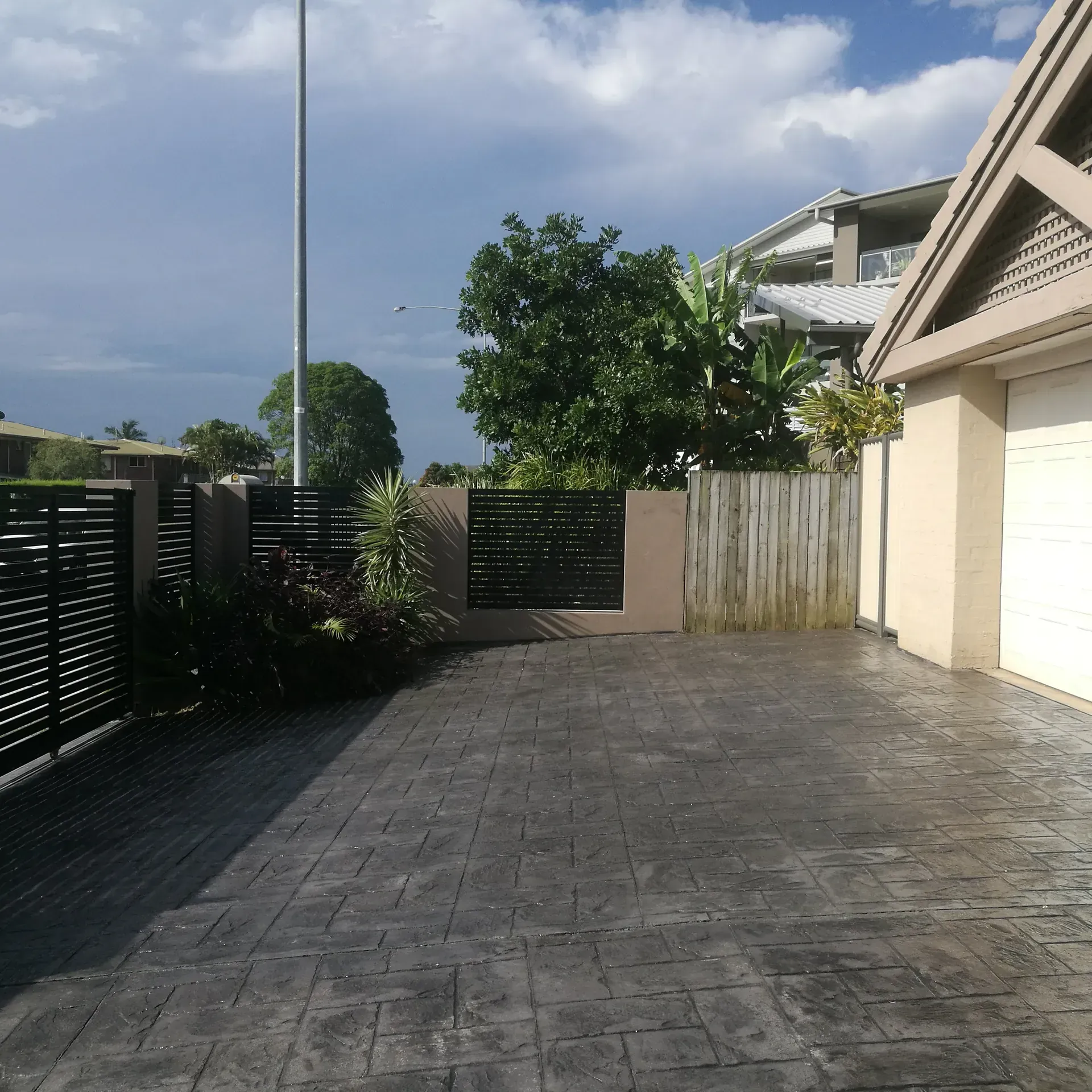 Driveway with black gate, concrete, beige garage, and cloudy sky.