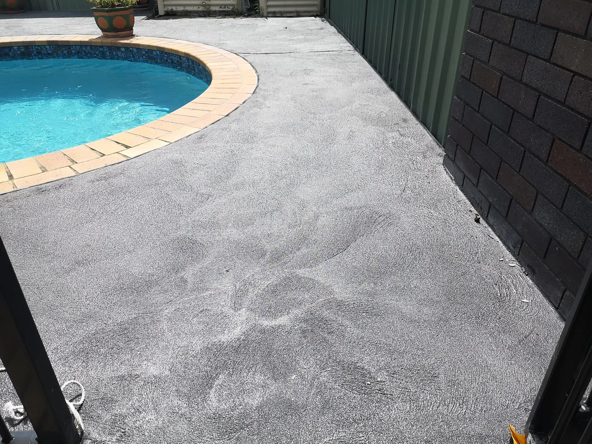 Poolside patio with gray speckled concrete, brick wall, and green fence. Pool has blue water and a brick border.