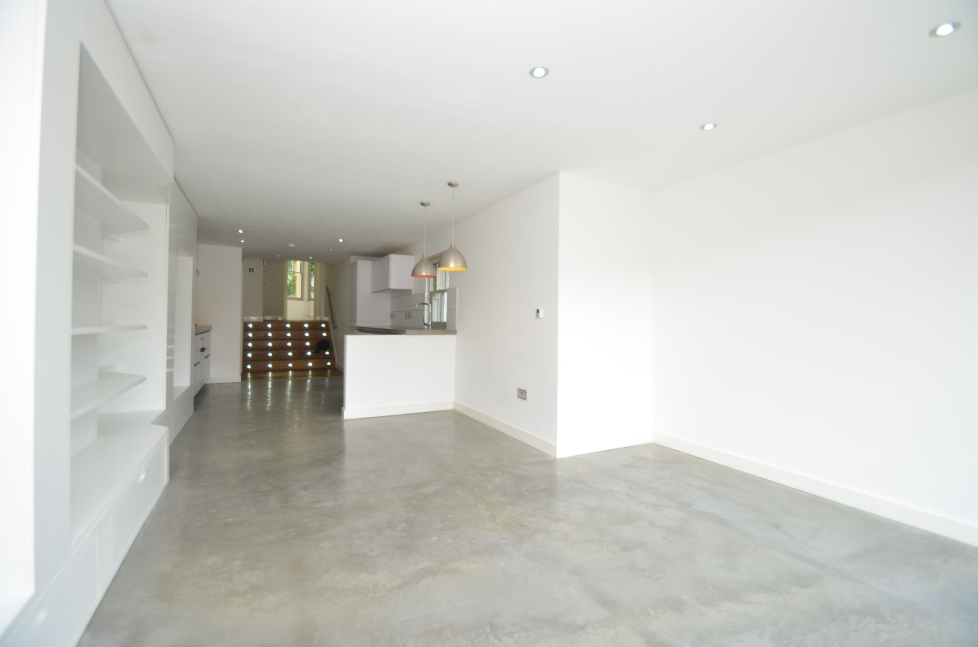 Empty, modern-style room with white walls, built-in shelves, and a concrete floor. Kitchen visible in the background.