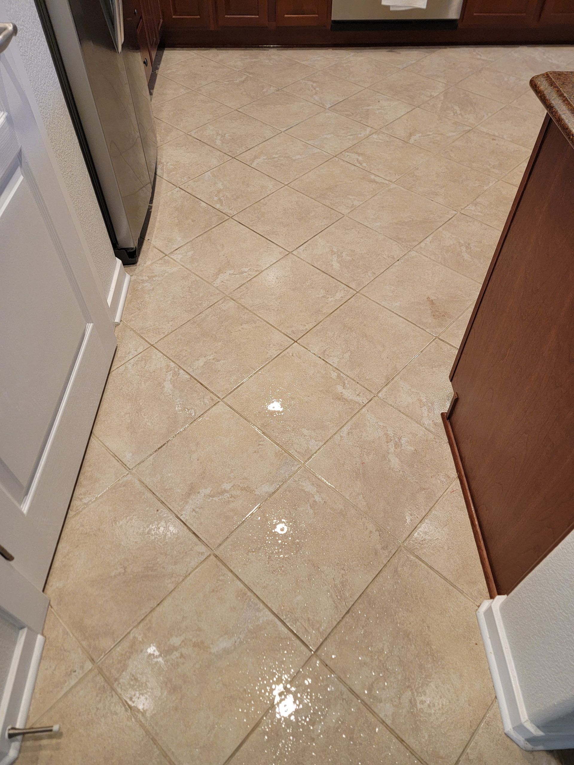 Kitchen floor with beige tile and water puddles near cabinets and a refrigerator