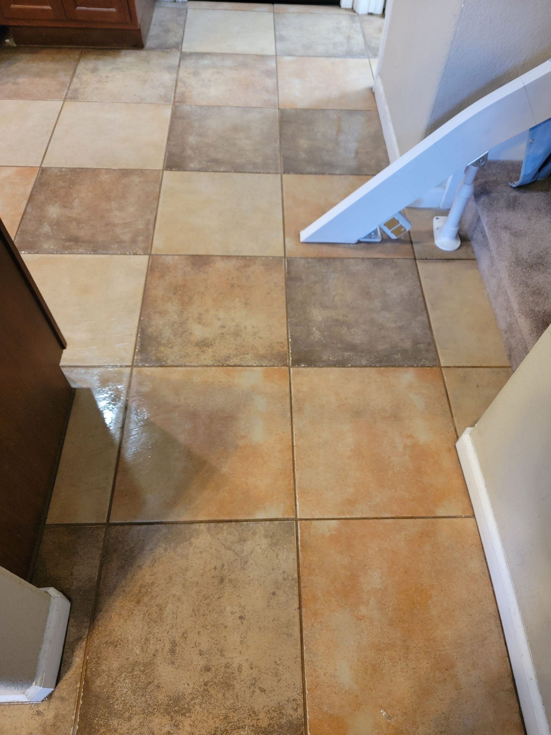 A view of a floor covered in square, mottled, earth-toned tiles in shades of beige, brown, and tan near carpeted stairs.