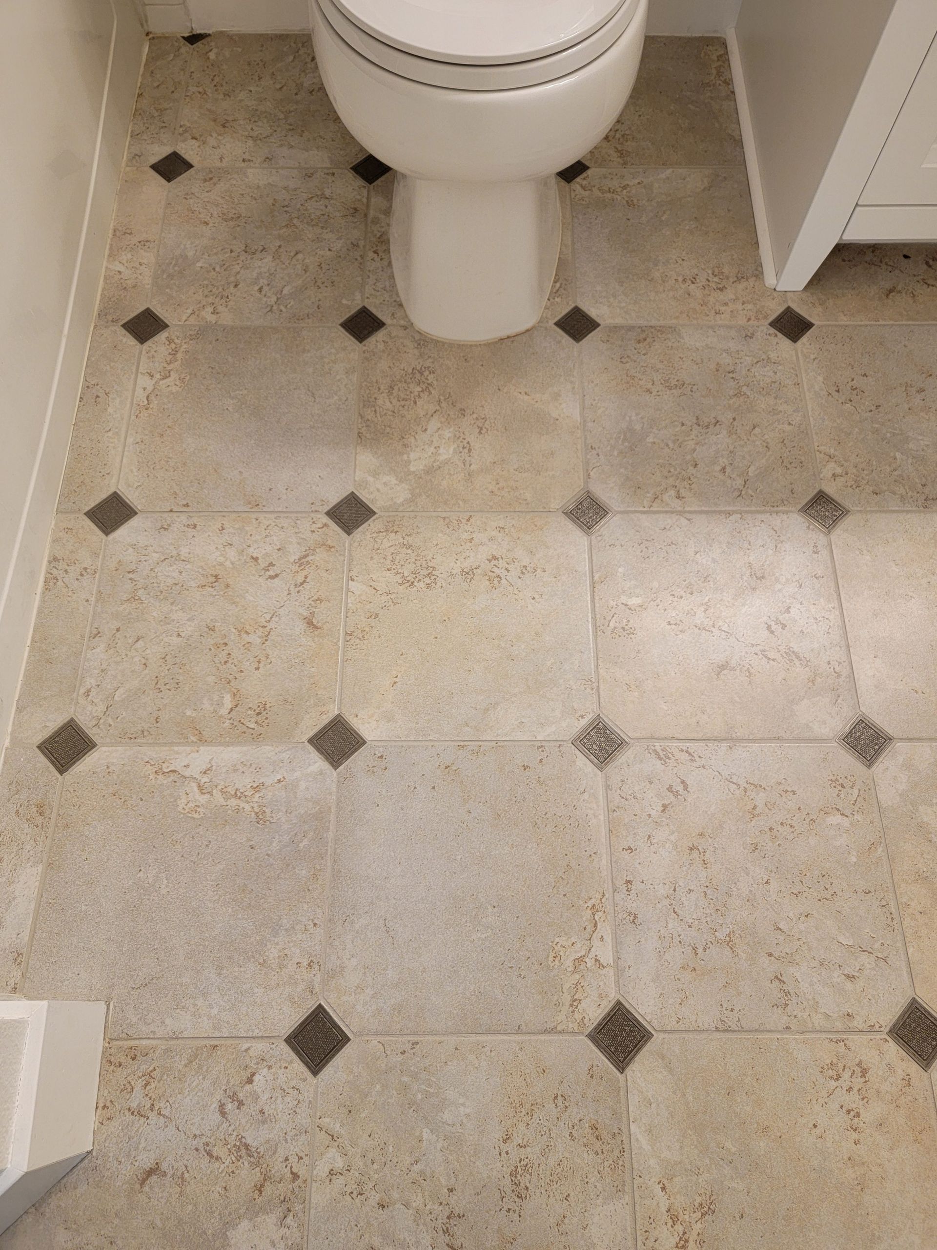 Bathroom floor featuring large square beige tiles accented by small, dark, square corner inserts.