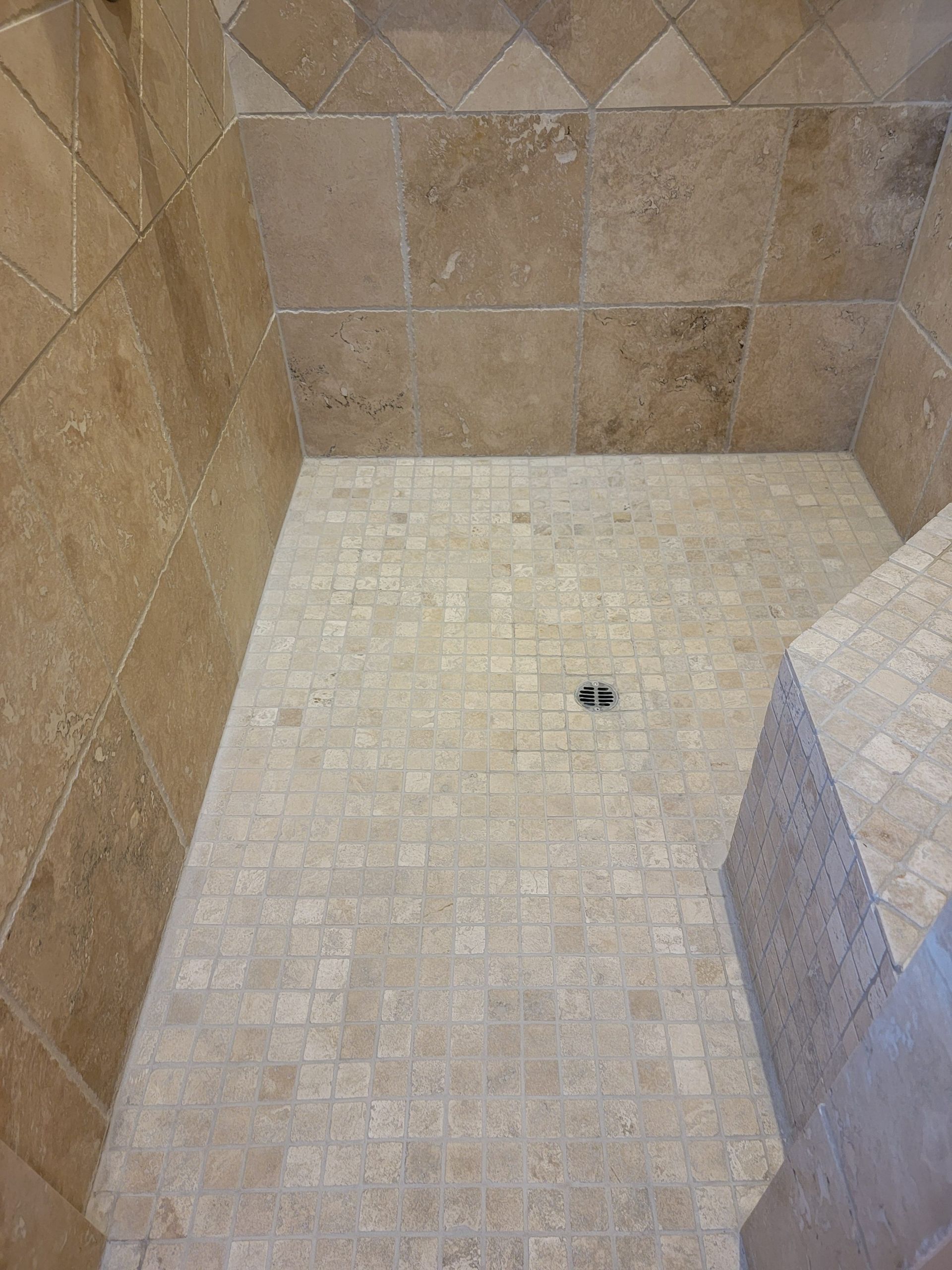 A walk-in shower with tan, square tile walls and a light-colored, small mosaic tile floor with a centered drain.
