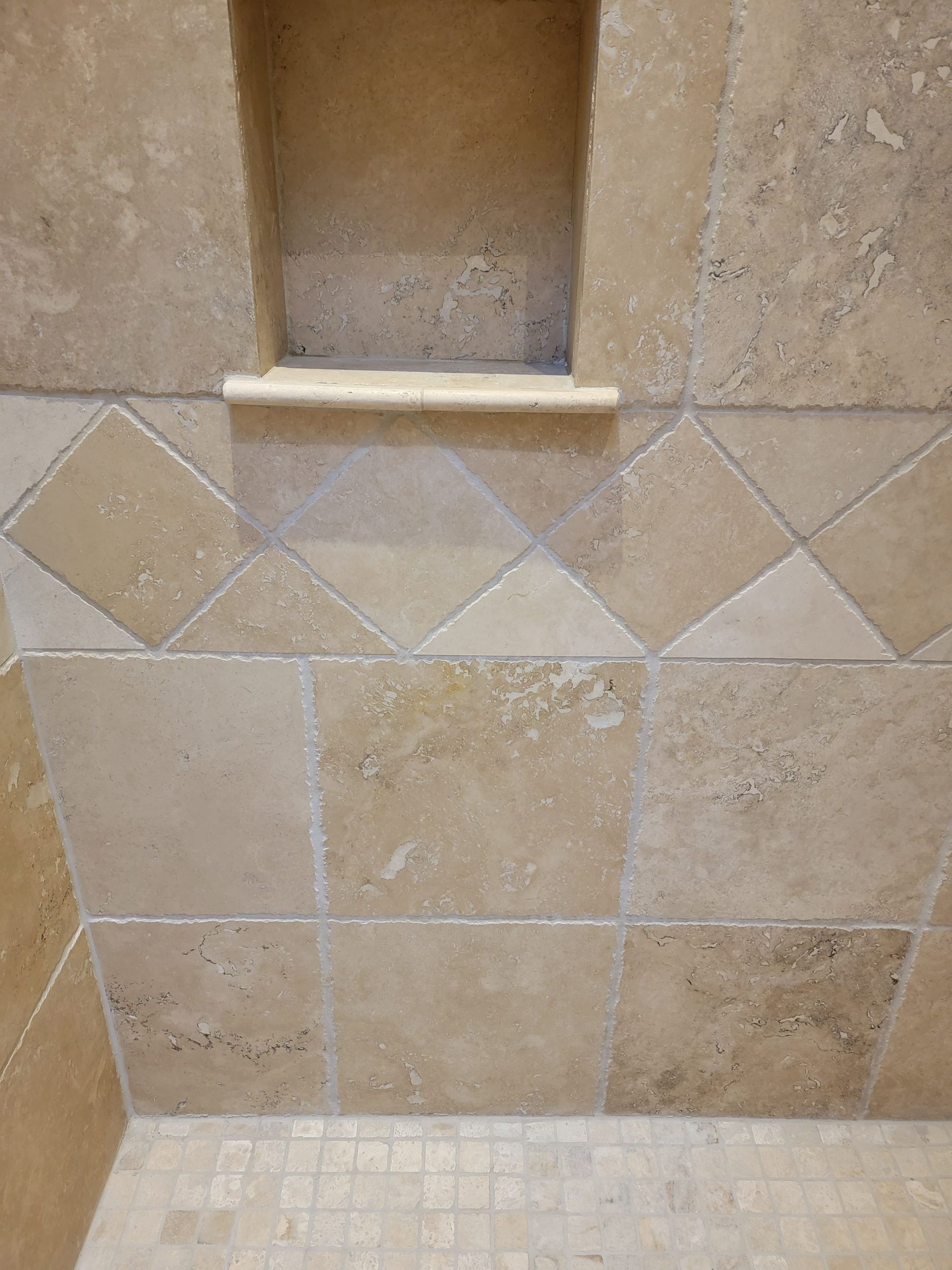 A tiled shower wall featuring tan travertine stone, a recessed niche, and a decorative border of diamond-patterned tiles.
