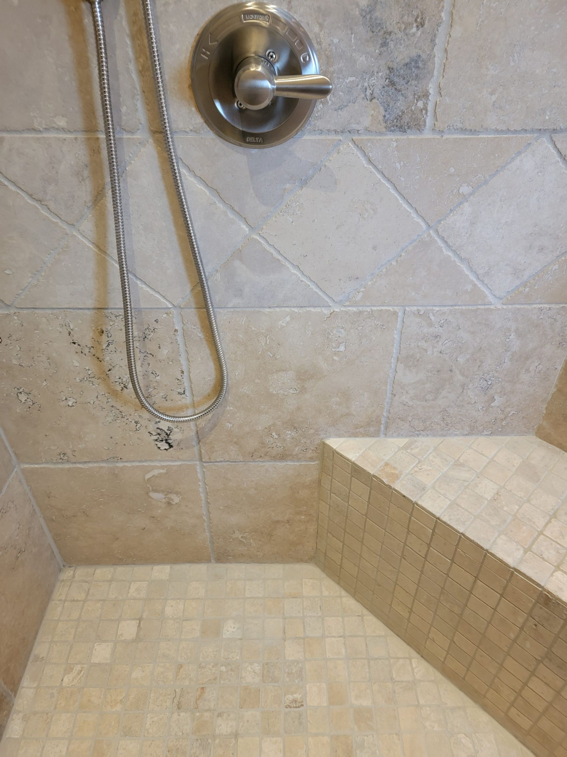 A shower stall with light beige stone tile walls and a built-in corner bench, featuring a metallic shower handle.