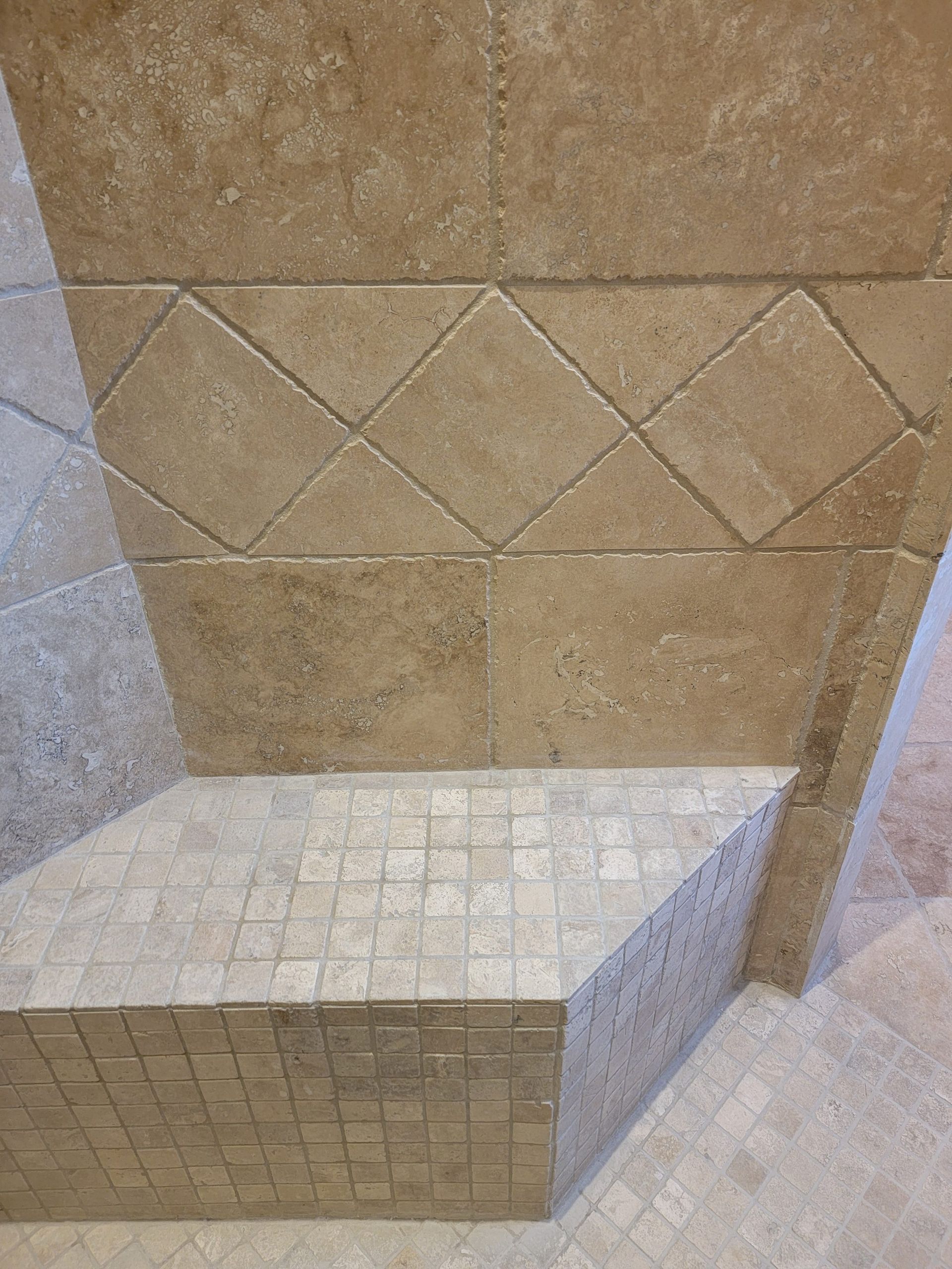 A corner shower bench tiled with small, light-toned stone squares, set against wall tiles in a diamond and block pattern.