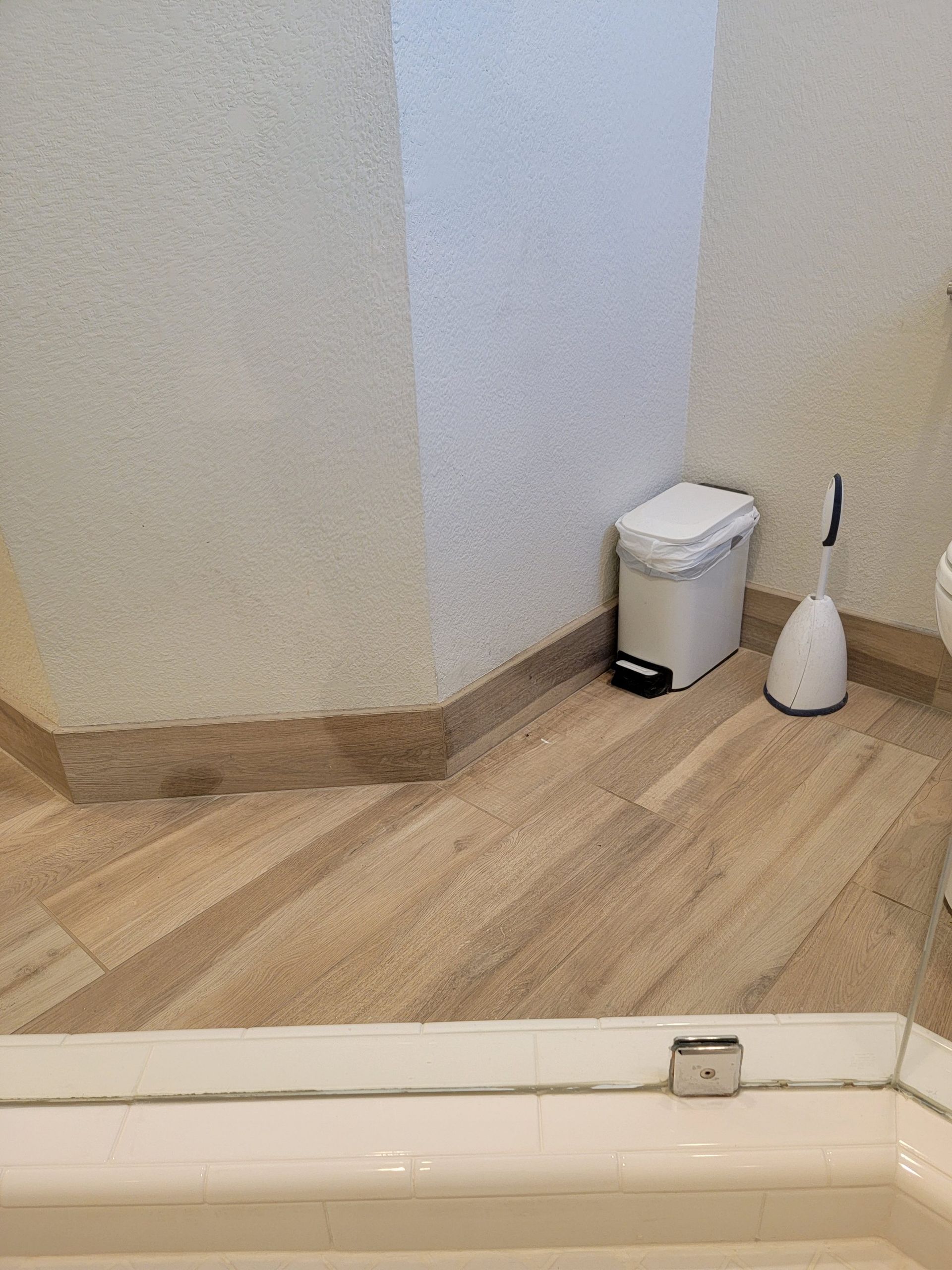 A white waste bin and a toilet brush stand in the corner of a tiled bathroom floor next to a glass shower enclosure.