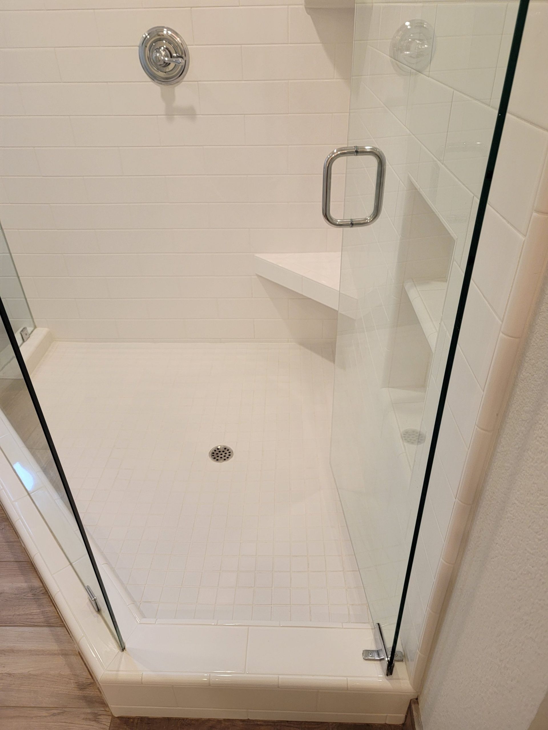 A white, corner walk-in shower with a glass door, built-in corner bench, white tile walls, and a drain in the floor.