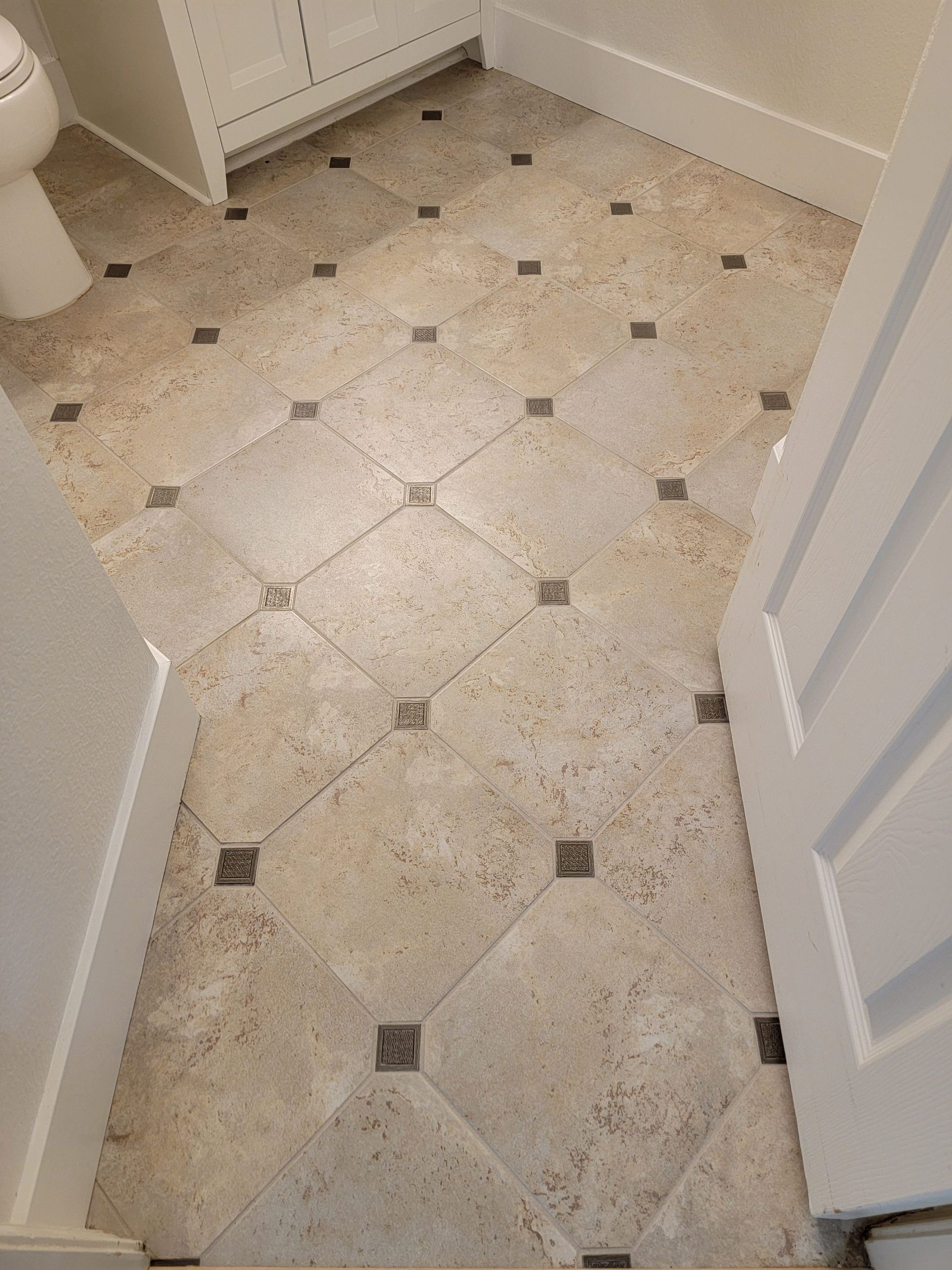 A bathroom floor featuring large, light beige square tiles set diagonally with small, dark square accent tiles at corners.