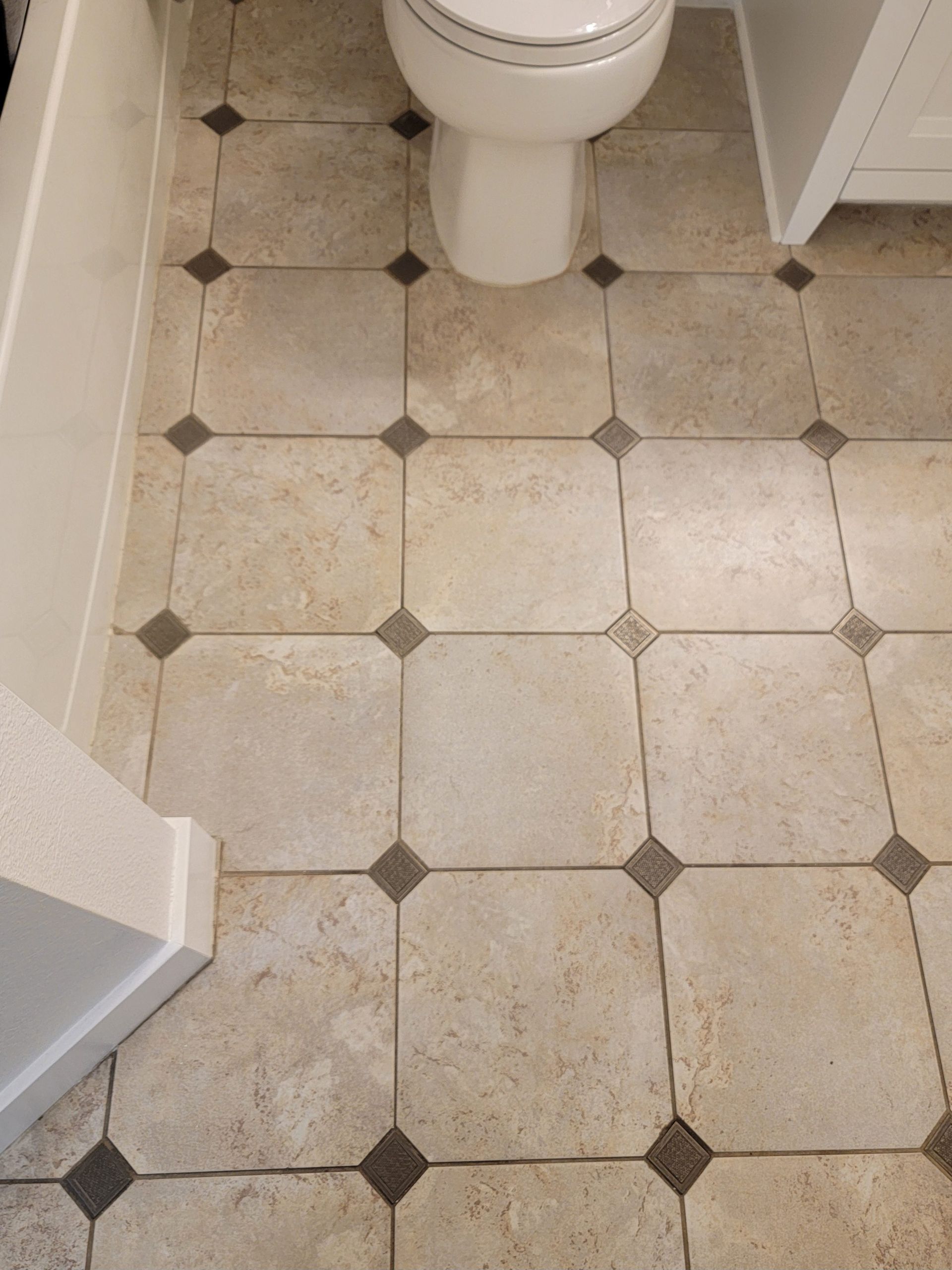 A bathroom floor featuring beige square tiles with small, dark square corner accents, surrounding a white toilet base.