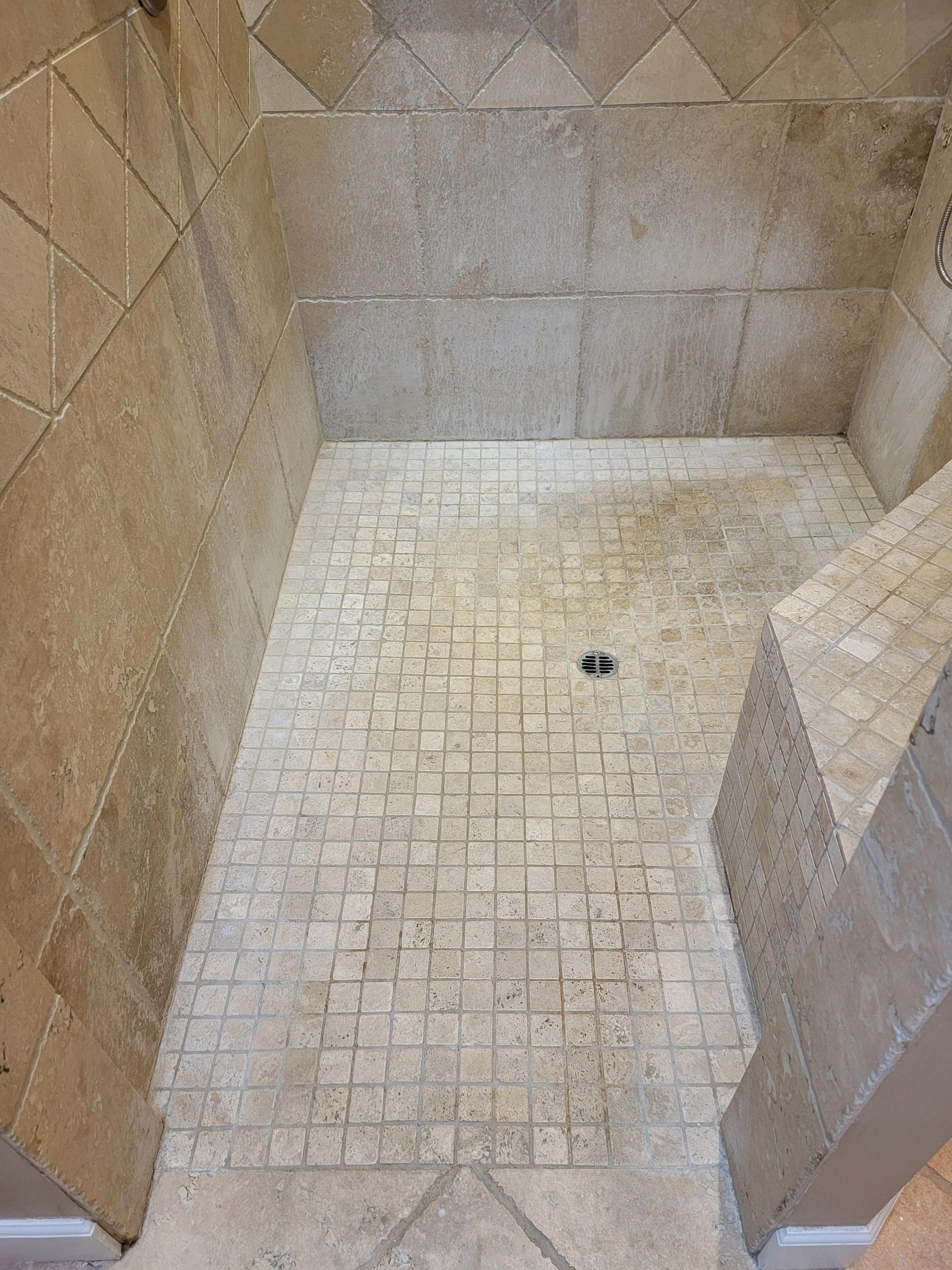 A walk-in shower with light tan stone tile walls and a mosaic stone floor featuring a centered drain.