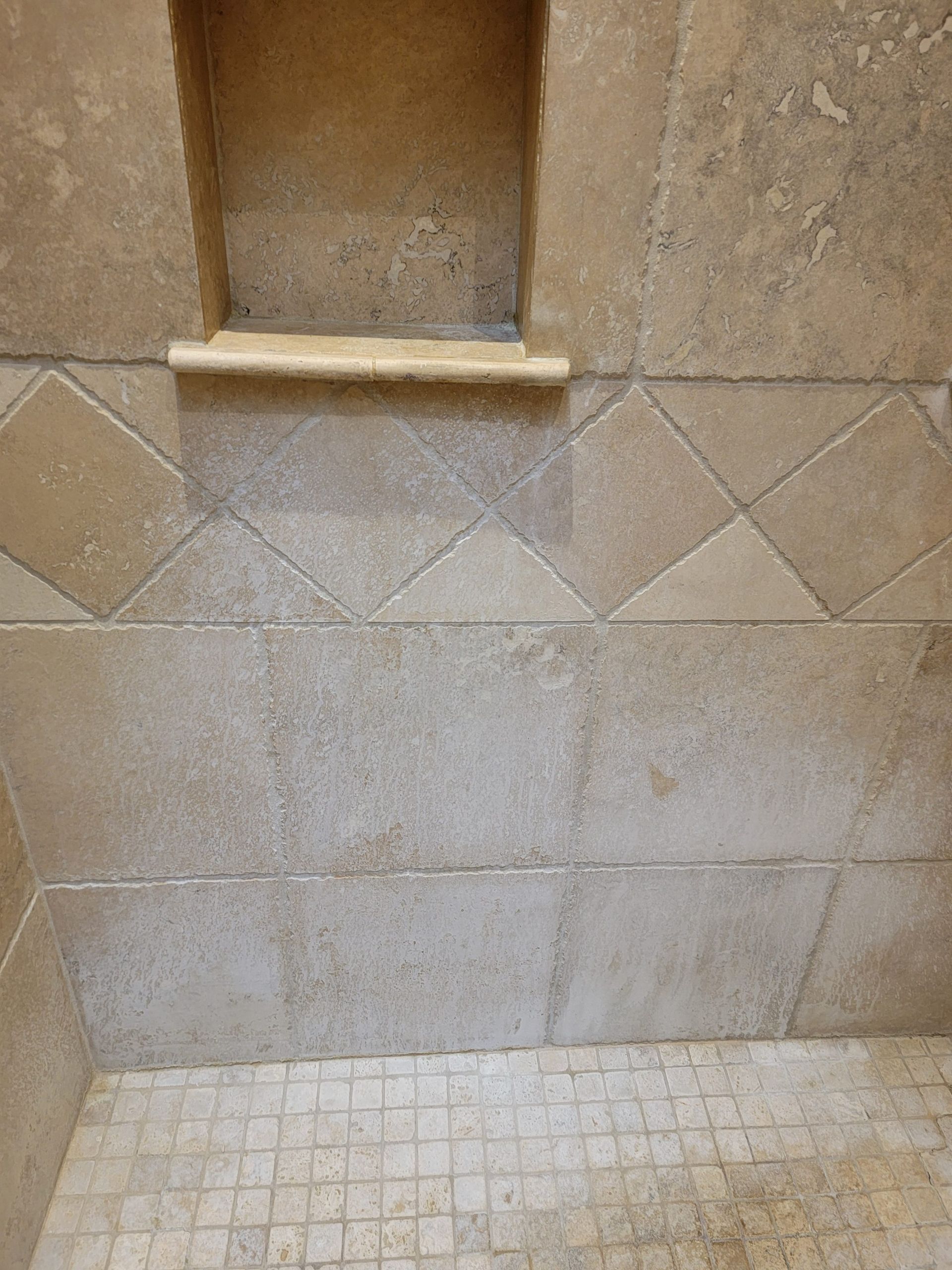 Tan natural stone shower wall with a square recessed niche and a mosaic tile floor showing signs of mineral buildup.
