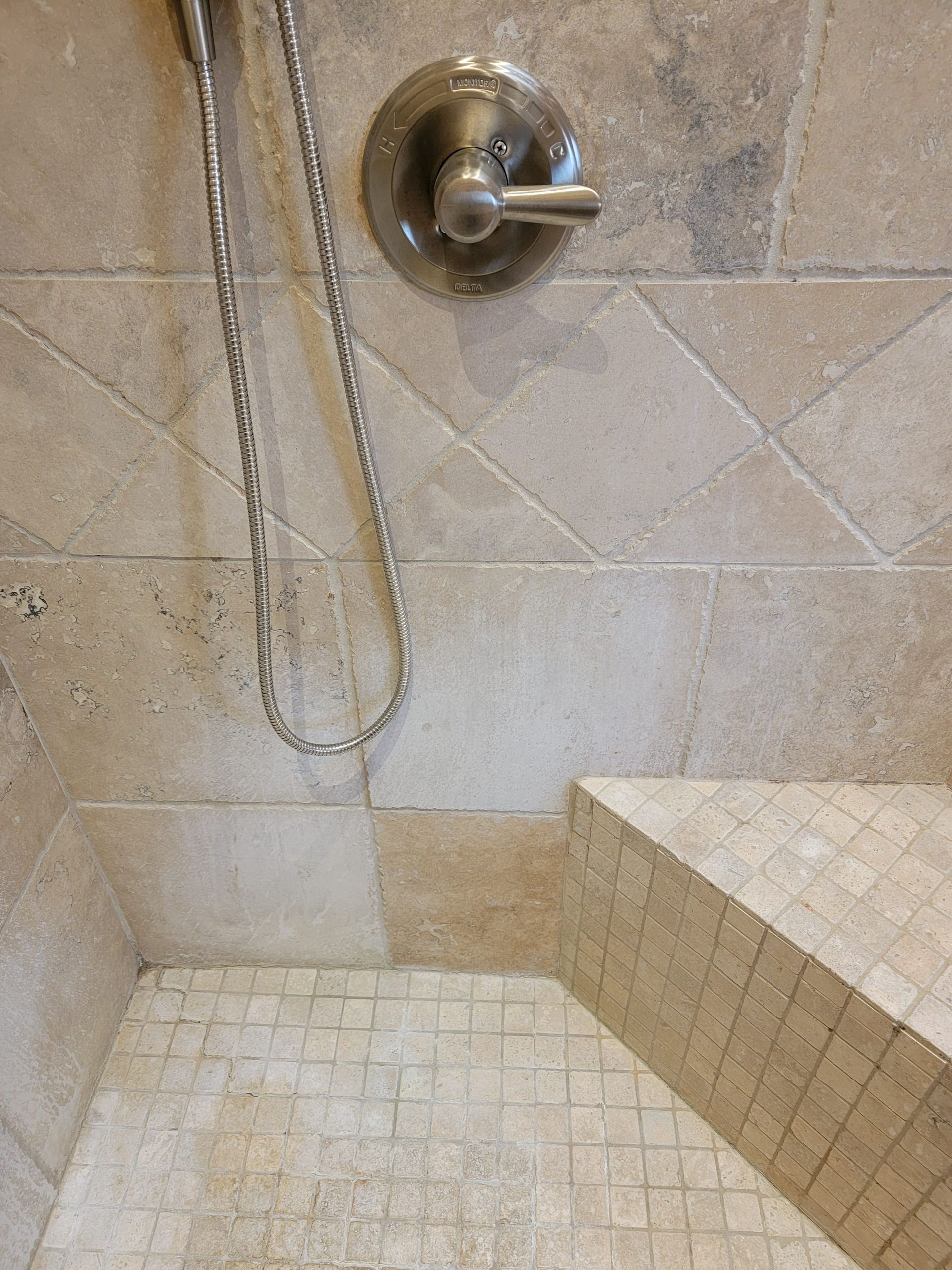 A tiled shower interior featuring a stone-colored wall, a chrome mixer valve, a hanging shower hose, and a corner bench.