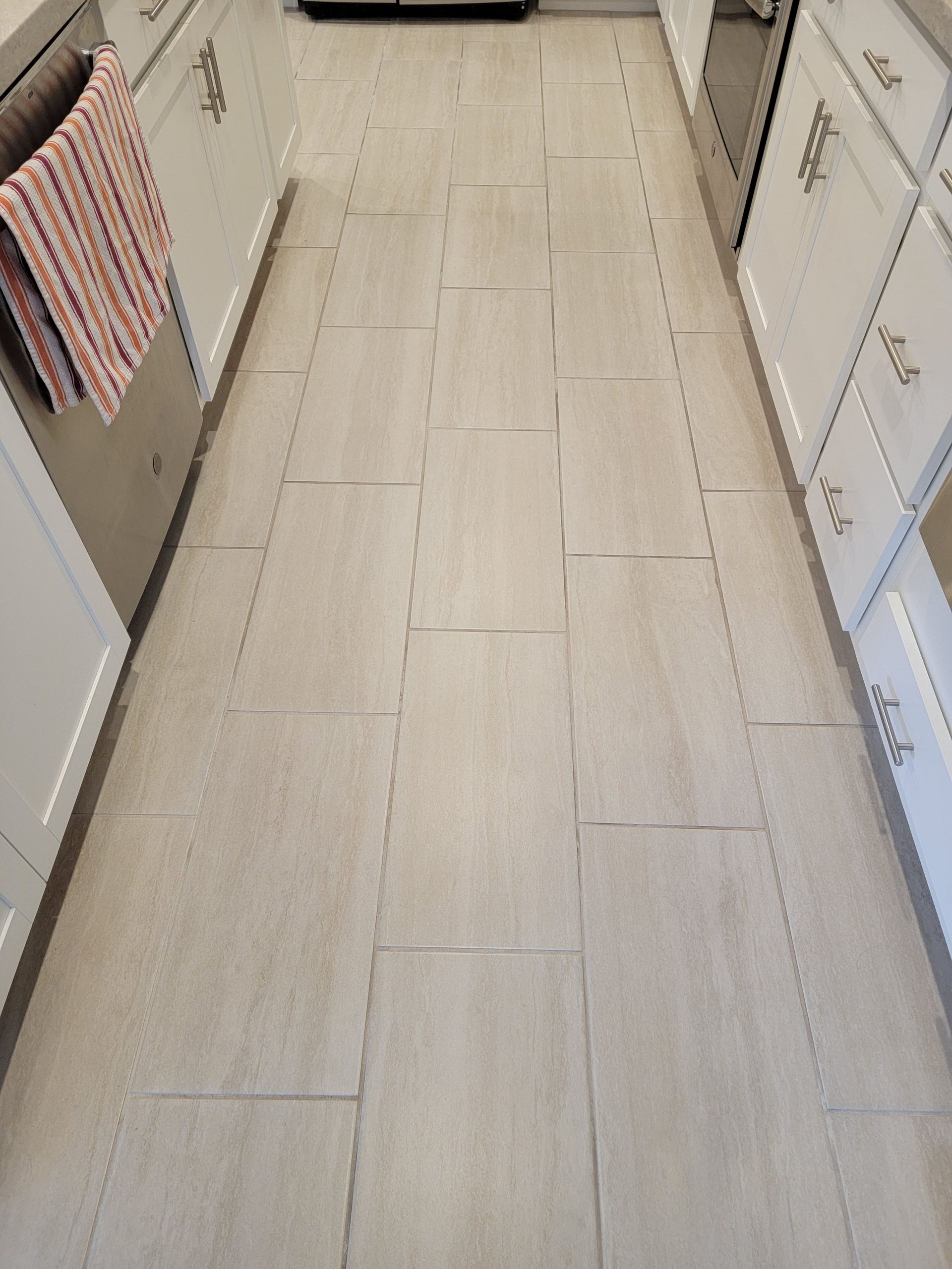 Light-colored wood-look ceramic floor tiles installed in a kitchen with white cabinetry and a patterned hand towel.