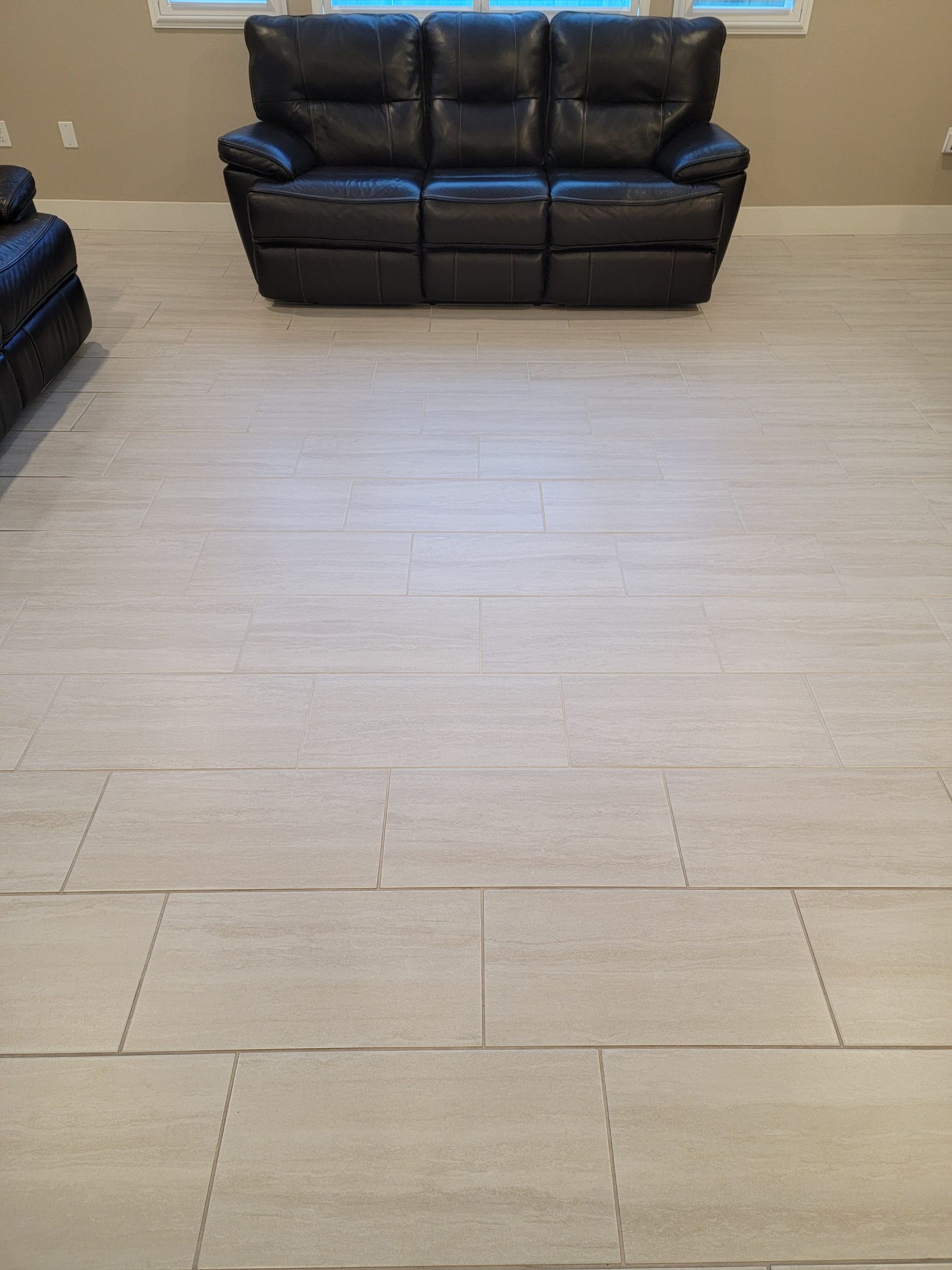 A black leather three-cushion sofa sits centered against a wall, facing a large room with light-colored tile flooring.