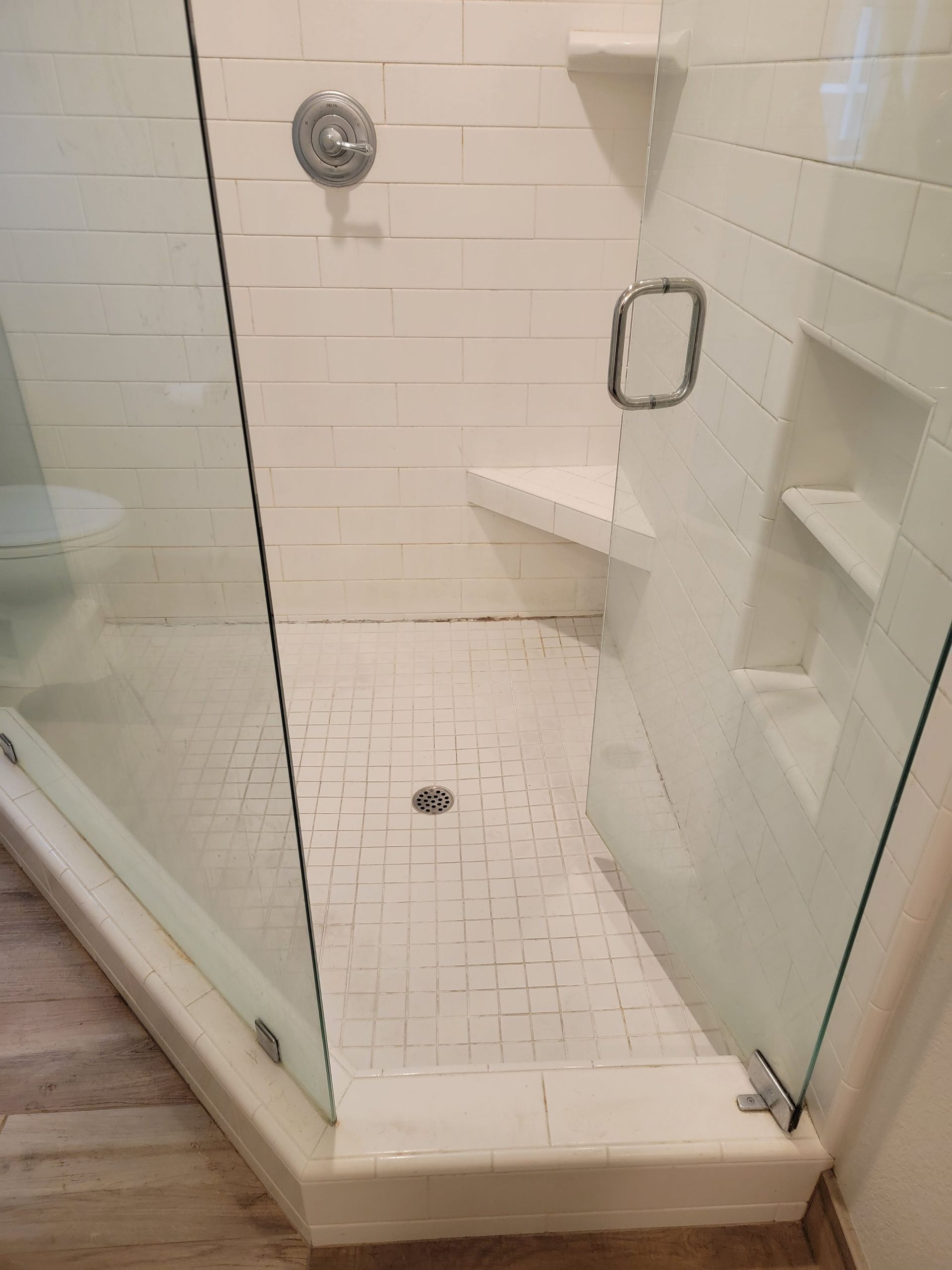 A modern shower enclosure with white tiled walls, a small built-in bench, a recessed shelf, and a glass door.