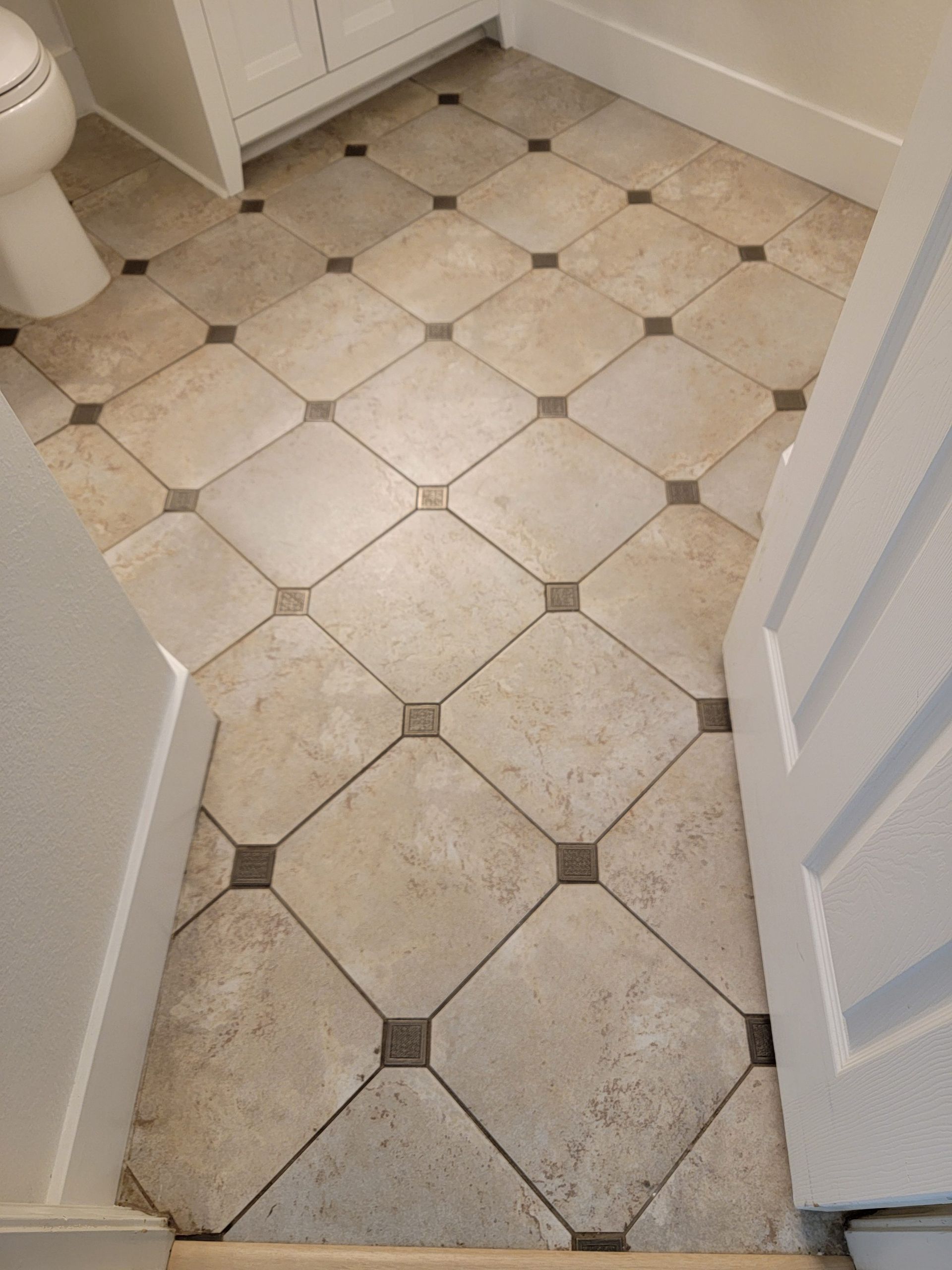 A high-angle view of a bathroom floor featuring beige square tiles laid diagonally with small dark square accent tiles.