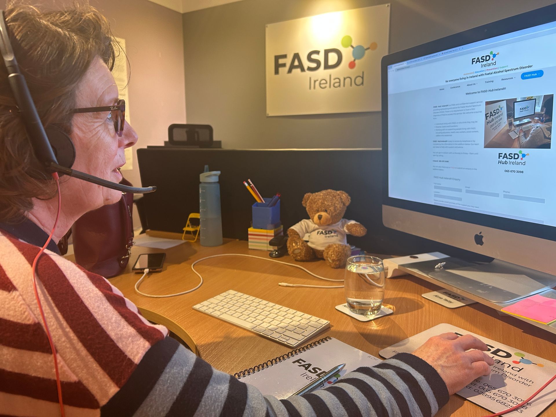 A female is sitting at a desk wearing a telephone headset is looking at a computer screen showing information about FASD Ireland