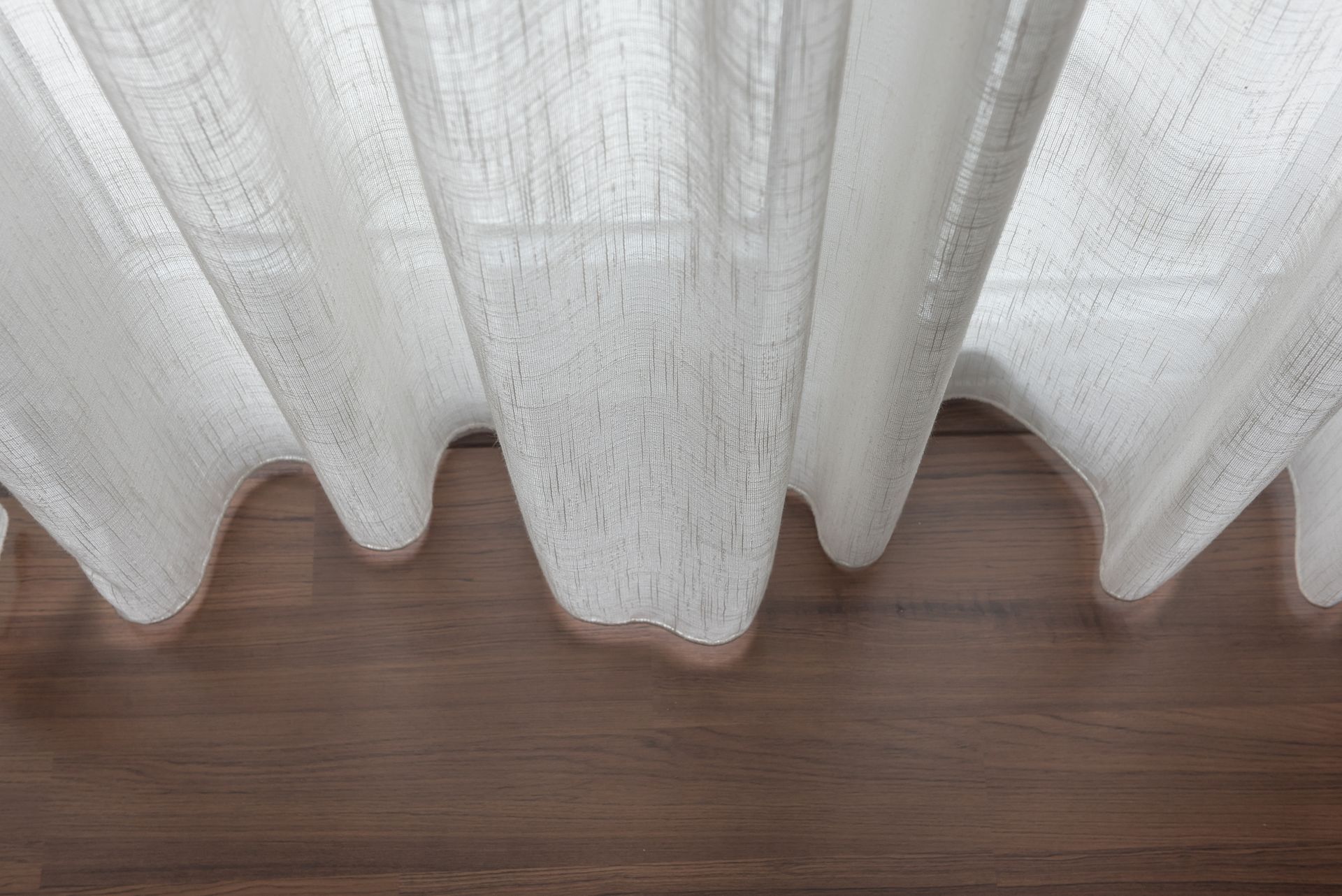 White sheer curtains hanging in front of a window, with wavy lower edge over a wooden floor.