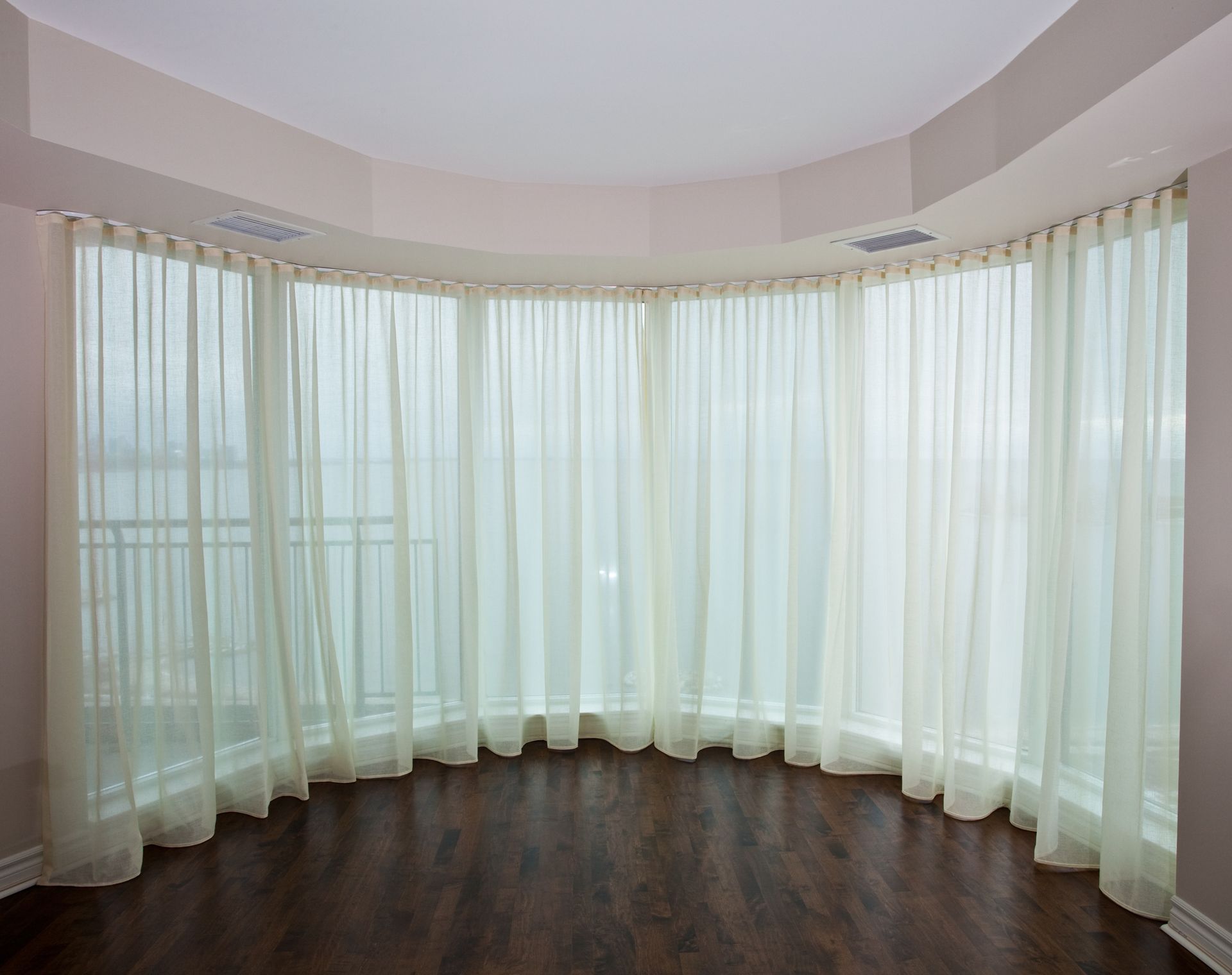 Curved window with sheer, cream-colored curtains and dark wood floor in an empty room.