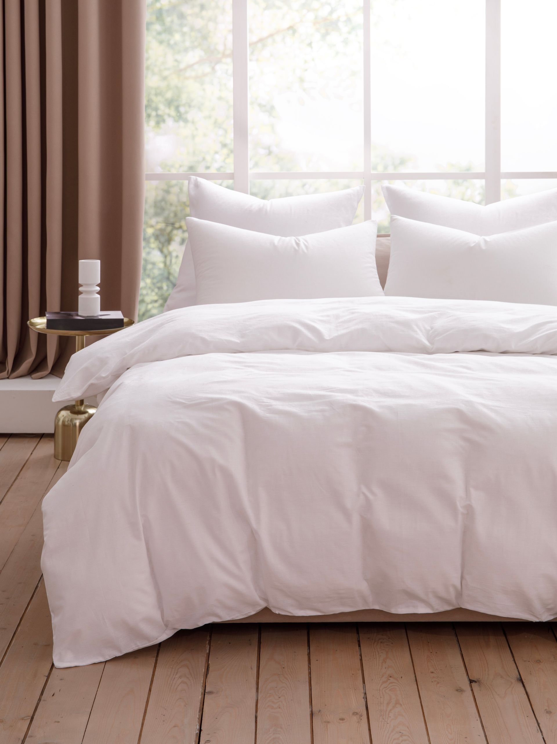 White bedding on a bed in a bright room with wood floors and a gold side table.
