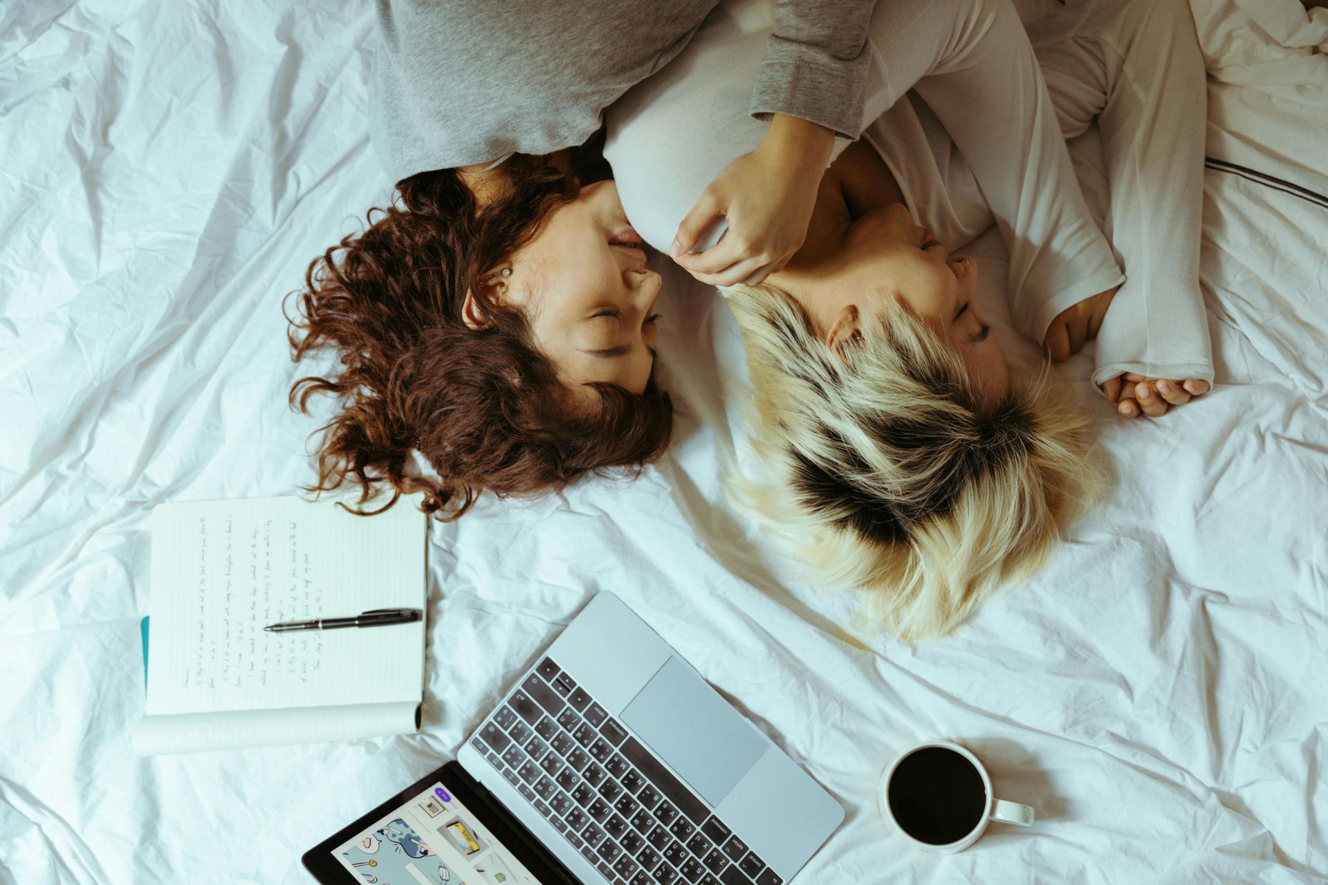 Two women are laying on a bed with a laptop and a cup of coffee.