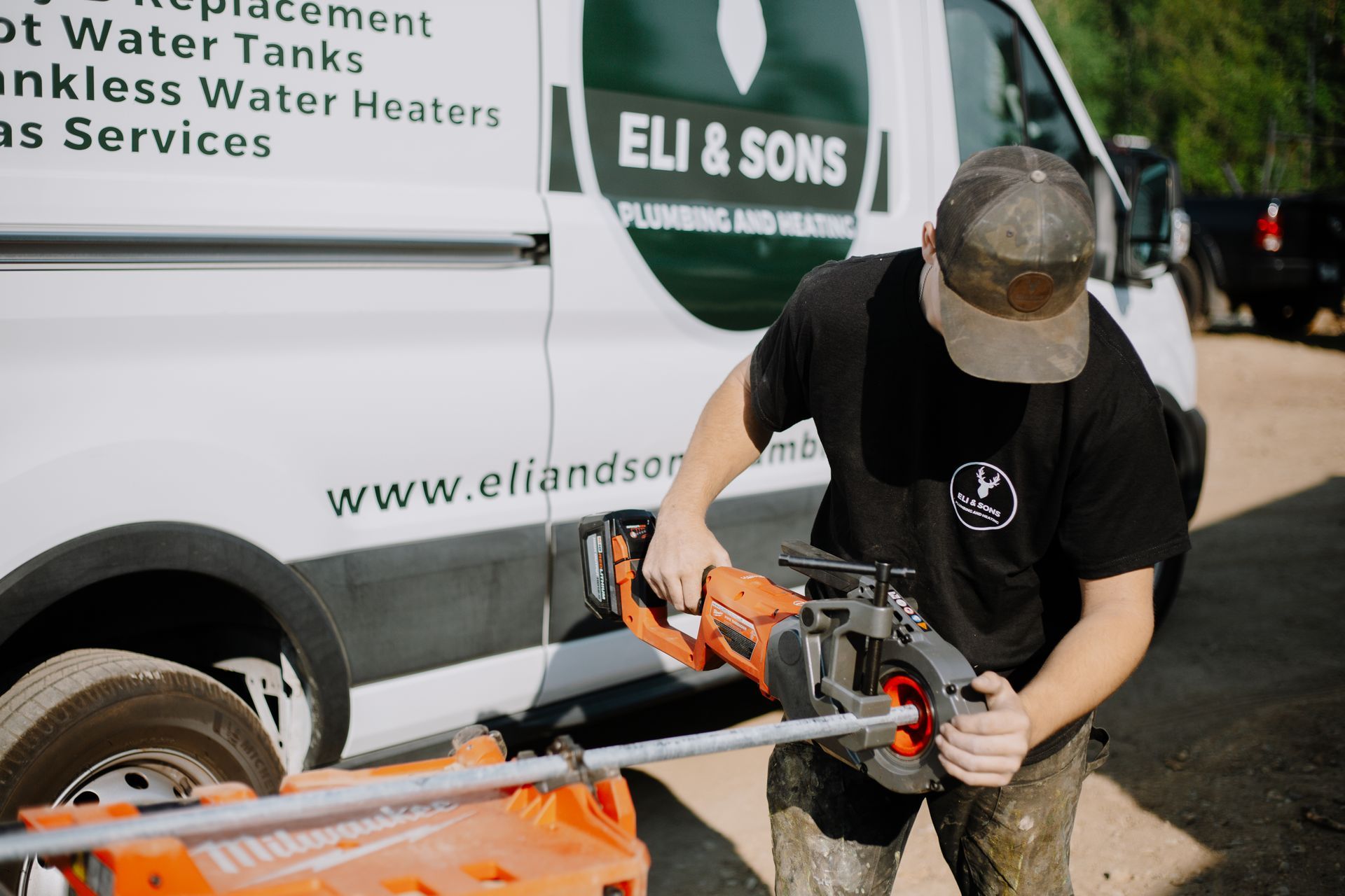 A man is working on a pipe in front of a van that says eli & sons