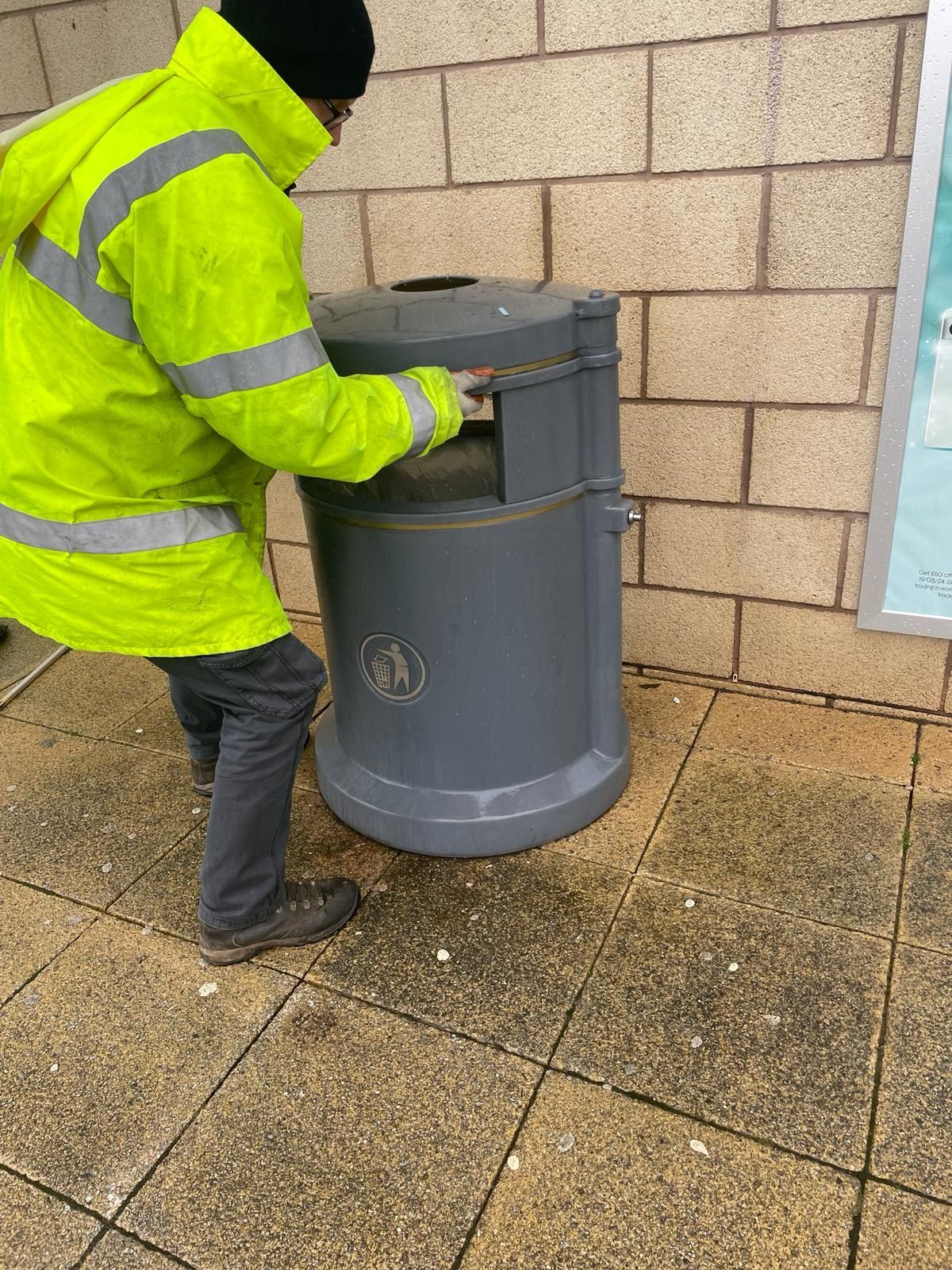 A man in a yellow jacket is standing next to a trash can.