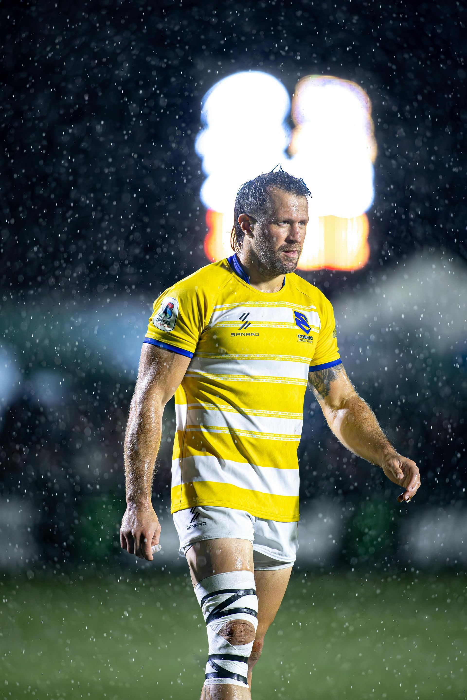 A man in a yellow and white striped shirt is standing in the rain on a soccer field.