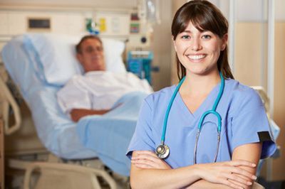A smiling nurse in blue scrubs with a stethoscope around their neck stands in front of a patient in a hospital bed.
