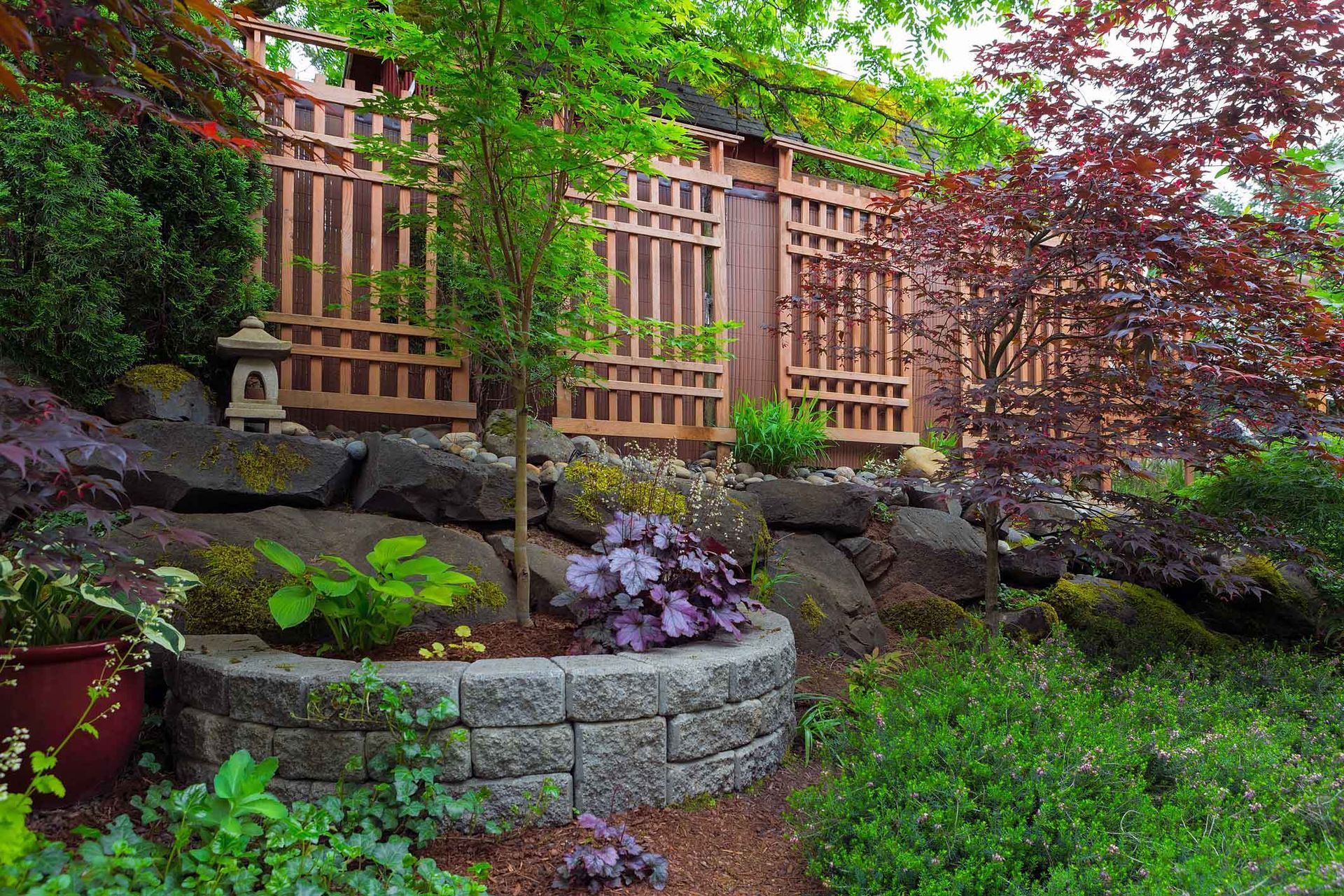 Japanese-inspired garden with tiered stone walls, wooden fence, and lush greenery.