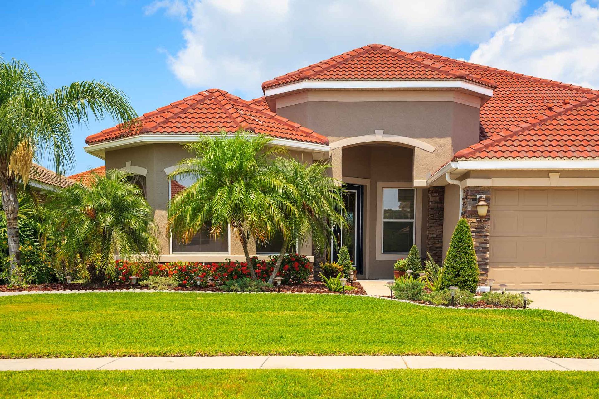 House with red tile roof, beige walls, palm trees, and green lawn under a blue sky.