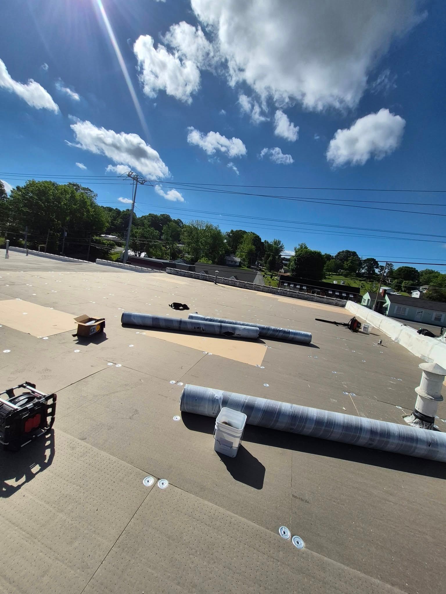 Rooftop with rolls of roofing material and equipment under a sunny blue sky with puffy clouds.