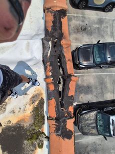 Aerial view of a damaged terracotta chimney. Black, deteriorated coating and holes are visible. Cars and a person are nearby.