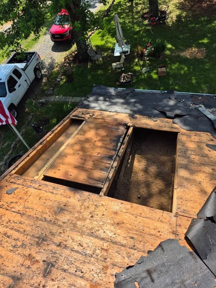 Overhead view of a damaged roof with large holes, revealing wood. A white truck and red car are parked nearby.